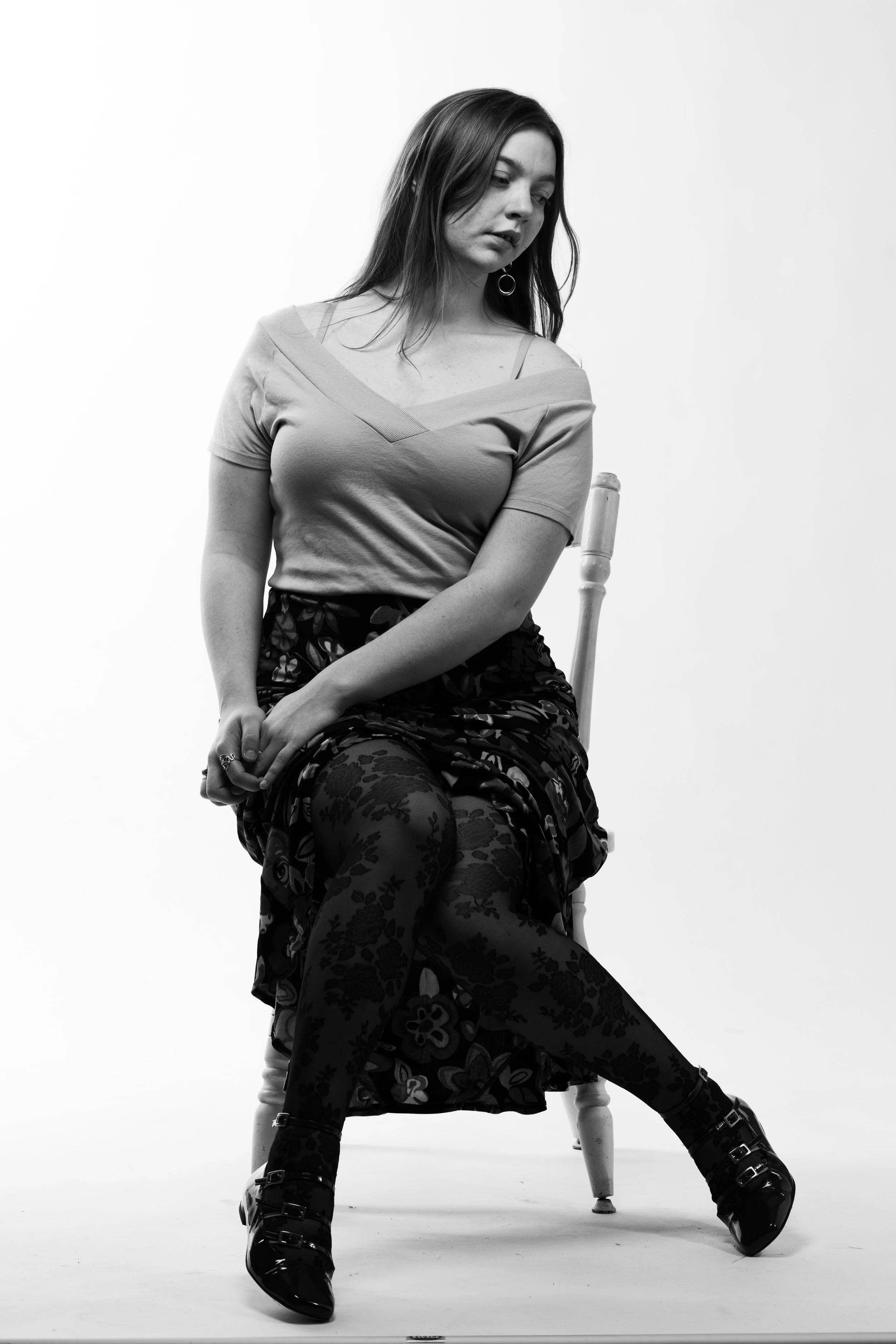 Black and white photo of a young woman sitting on a chair, wearing a top, patterned skirt, and high-heeled boots, with a contemplative expression.