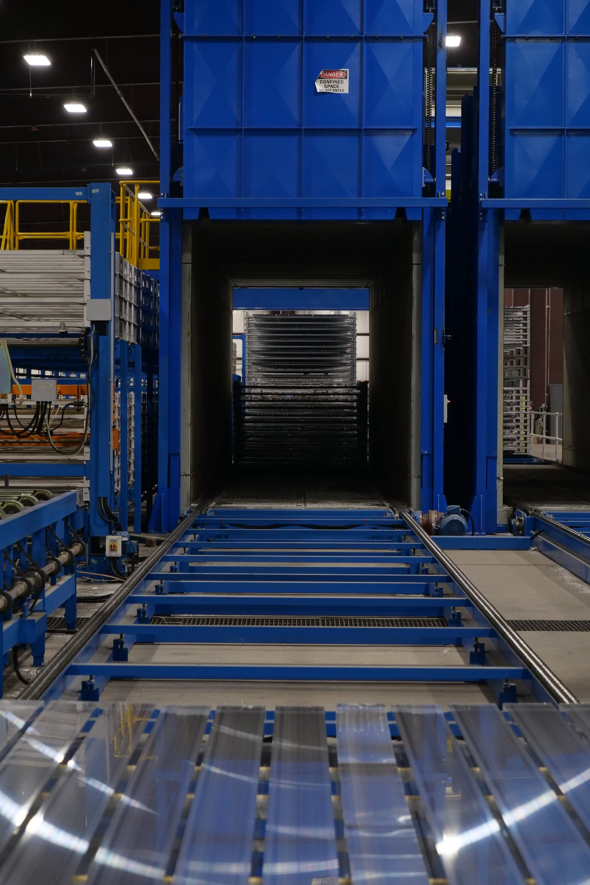 Inside an industrial warehouse with blue machinery and conveyor belt systems, with a warning sign that reads 'Danger: Confined Space Do Not Enter'.