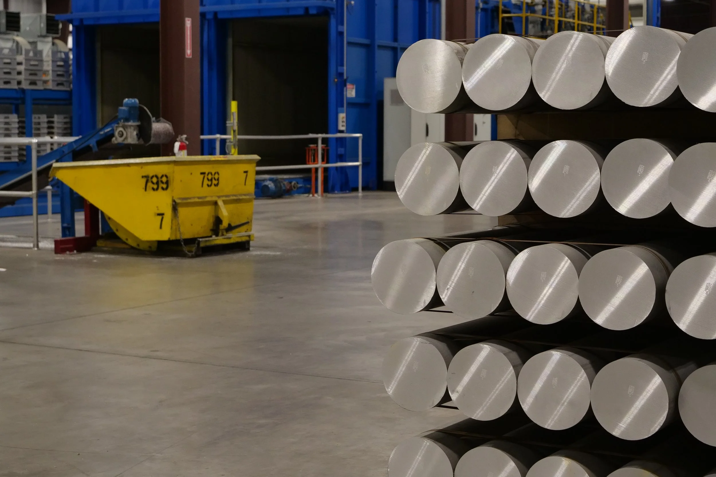 Industrial warehouse with large metal rolls stacked horizontally in the foreground and blue machinery and structures in the background.