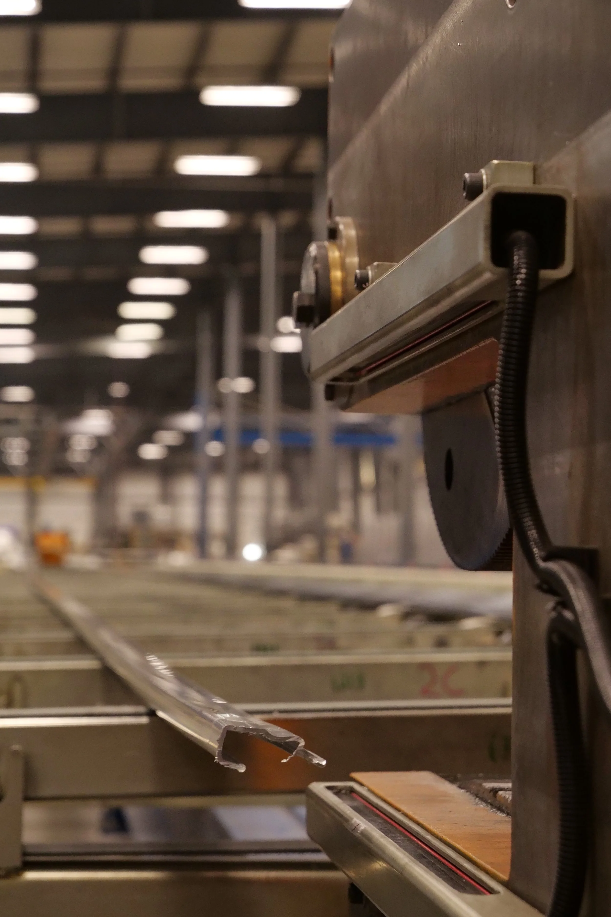 Close-up of a metal conveyor or machine part in an industrial or manufacturing setting, with blurred background of pipes, rails, and equipment.
