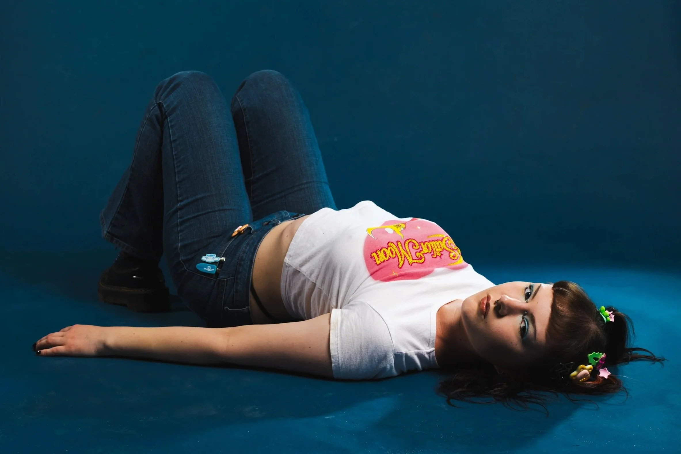 A woman lying on a dark blue floor with her head turned to the side, wearing a white t-shirt with pink and yellow text, blue jeans, and decorated with colorful hair accessories. The background is a solid dark blue color.