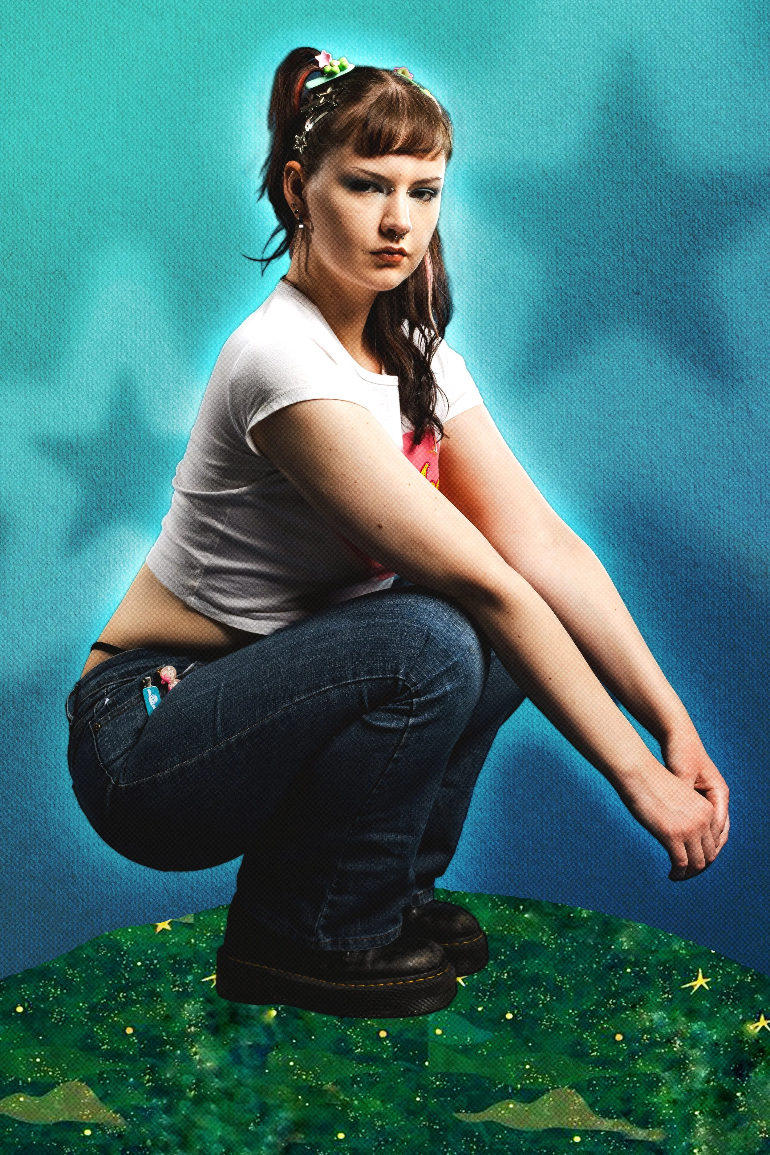 A young woman with red hair, large earrings, and multiple hair clips is squatting on a green, star-dotted surface with mountainous scenery in the background. She wears a white T-shirt, dark jeans, and black boots, with a serious expression on her fac