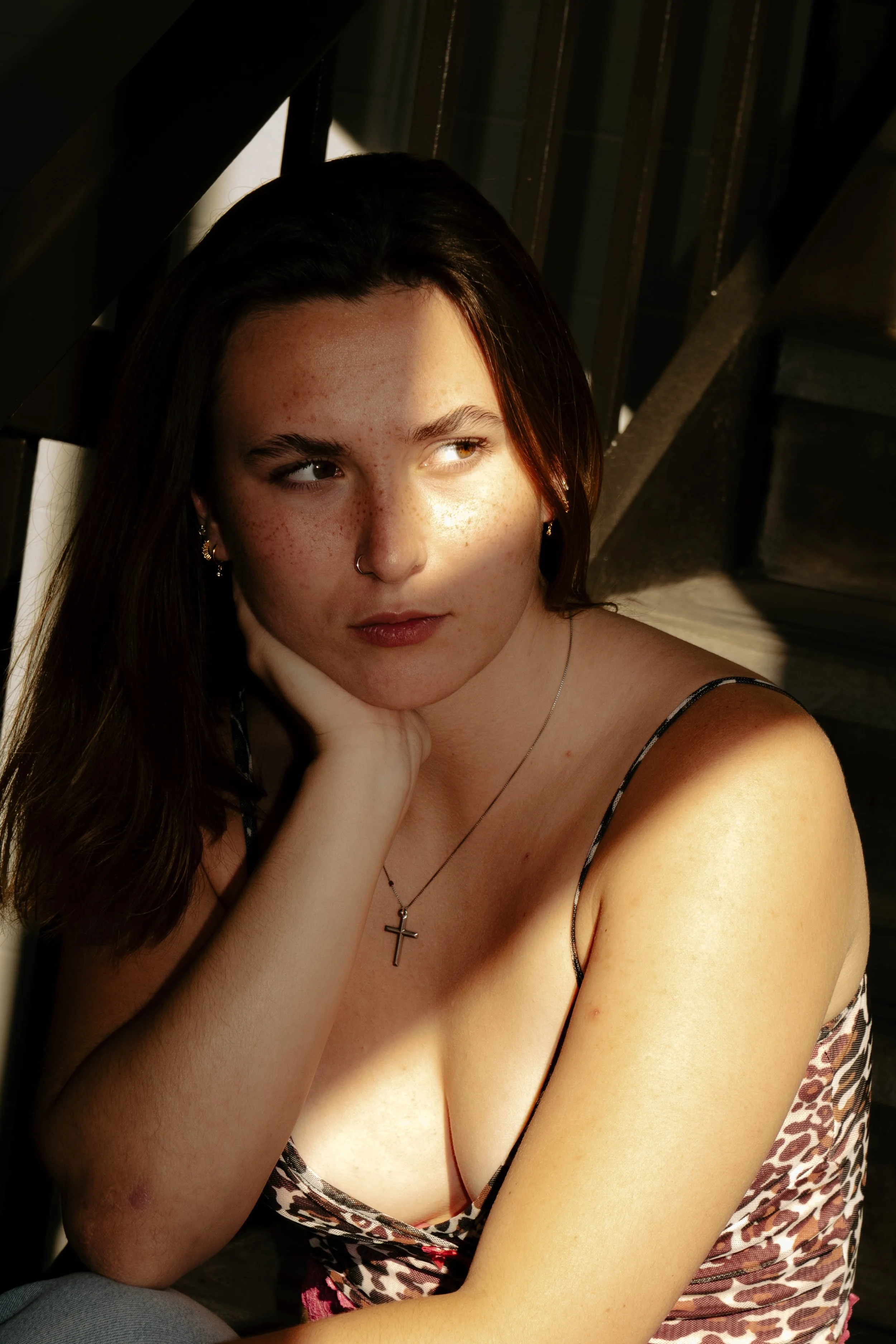 A young woman with dark hair and freckles, wearing a leopard print dress and a cross necklace, sits with her hand resting on her chin, illuminated by sunlight.