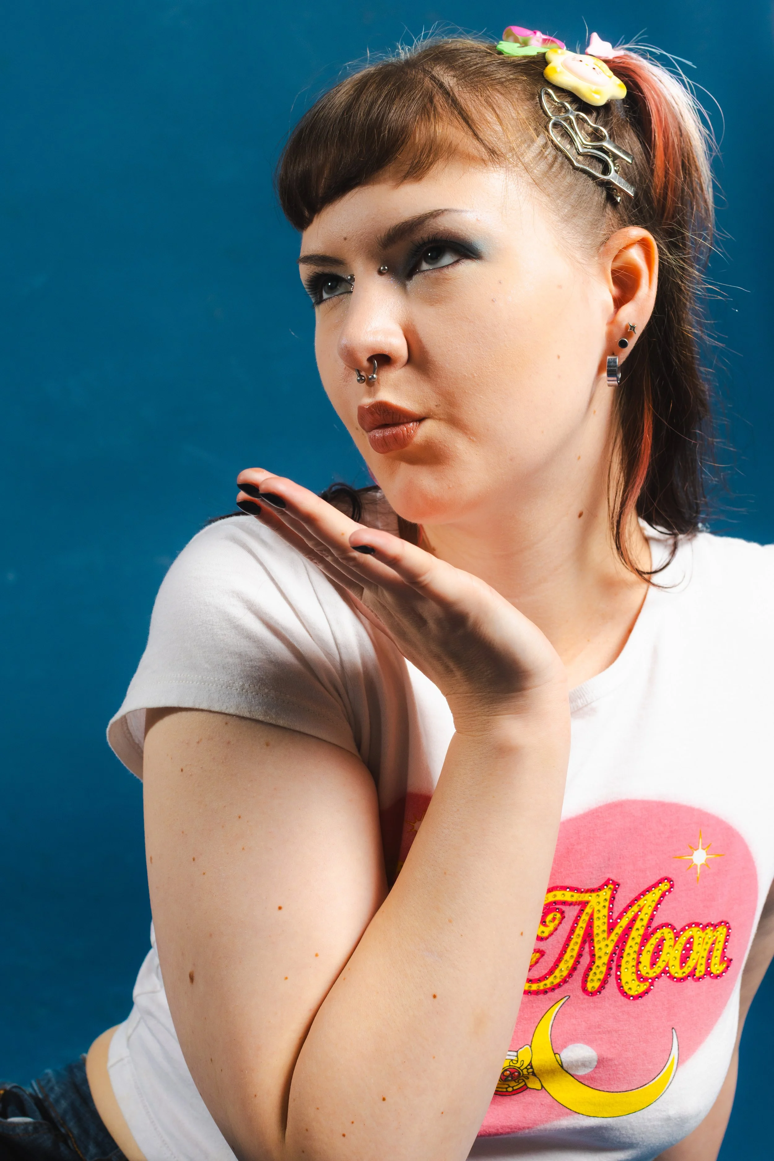 A woman with short brown hair and multiple earrings, including a septum piercing, posing with her hand under her chin against a blue background. She wears a white T-shirt with pink and yellow lettering and illustrations.