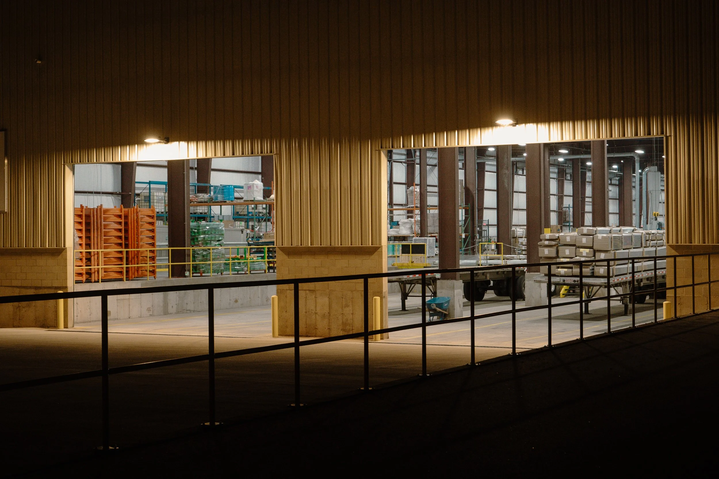 View of an indoor warehouse through a large window, showing stacked pallets, boxes, and shelves inside, illuminated by ceiling lights.