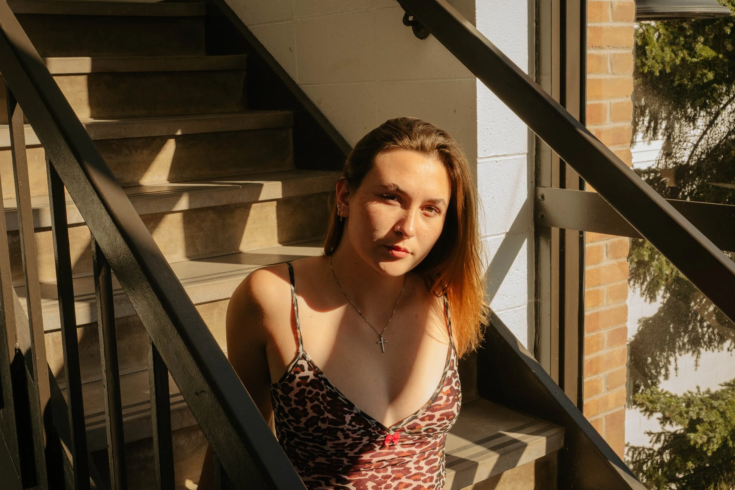 A young woman with long reddish hair sitting on outdoor stairs, wearing a leopard print dress, a cross necklace, and earrings, with sunlight casting shadows and trees visible in the background.