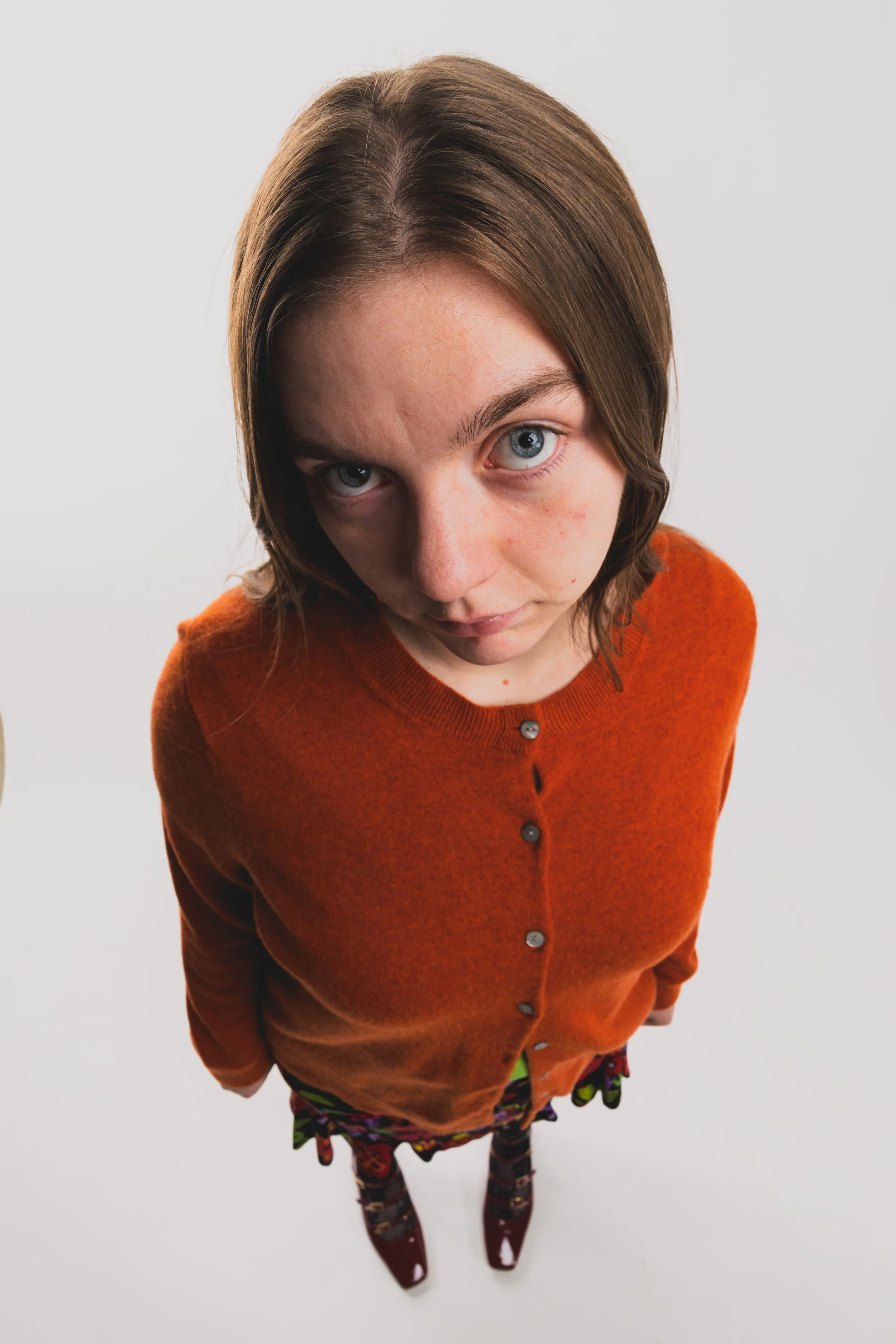 A young girl with shoulder-length brown hair and blue eyes, looking directly at the camera, wearing an orange cardigan with buttons over a colorful dress with pattern, and patterned tights.