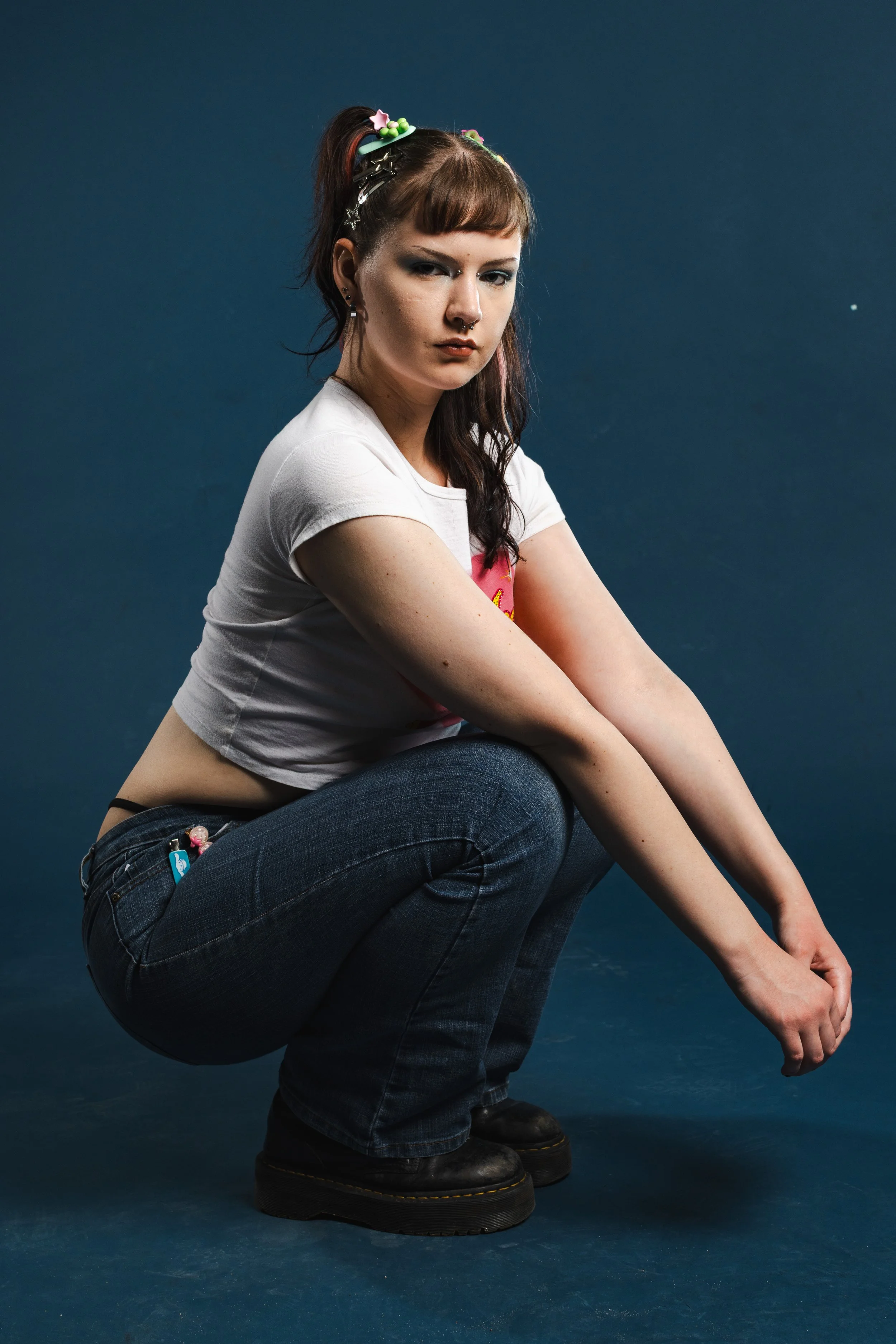 A young woman with brown hair styled in pigtails secured with colorful hair clips, wearing a white t-shirt and dark jeans, squatting on a blue floor with a dark blue background.