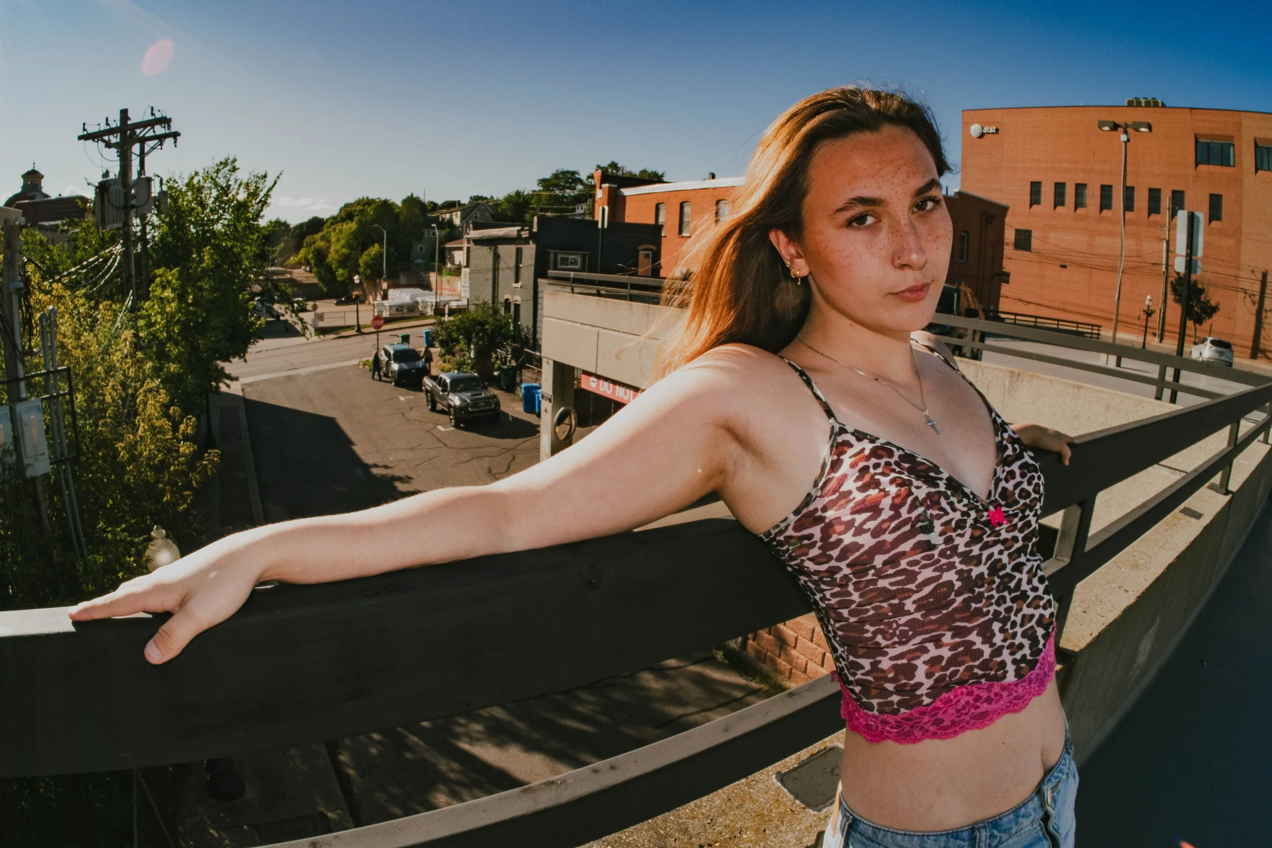 A young woman with light skin, freckles, and long brown hair taking a selfie on a rooftop balcony in an urban area, wearing a leopard print top and denim shorts, with a background of trees, parked cars, and building rooftops on a sunny day.