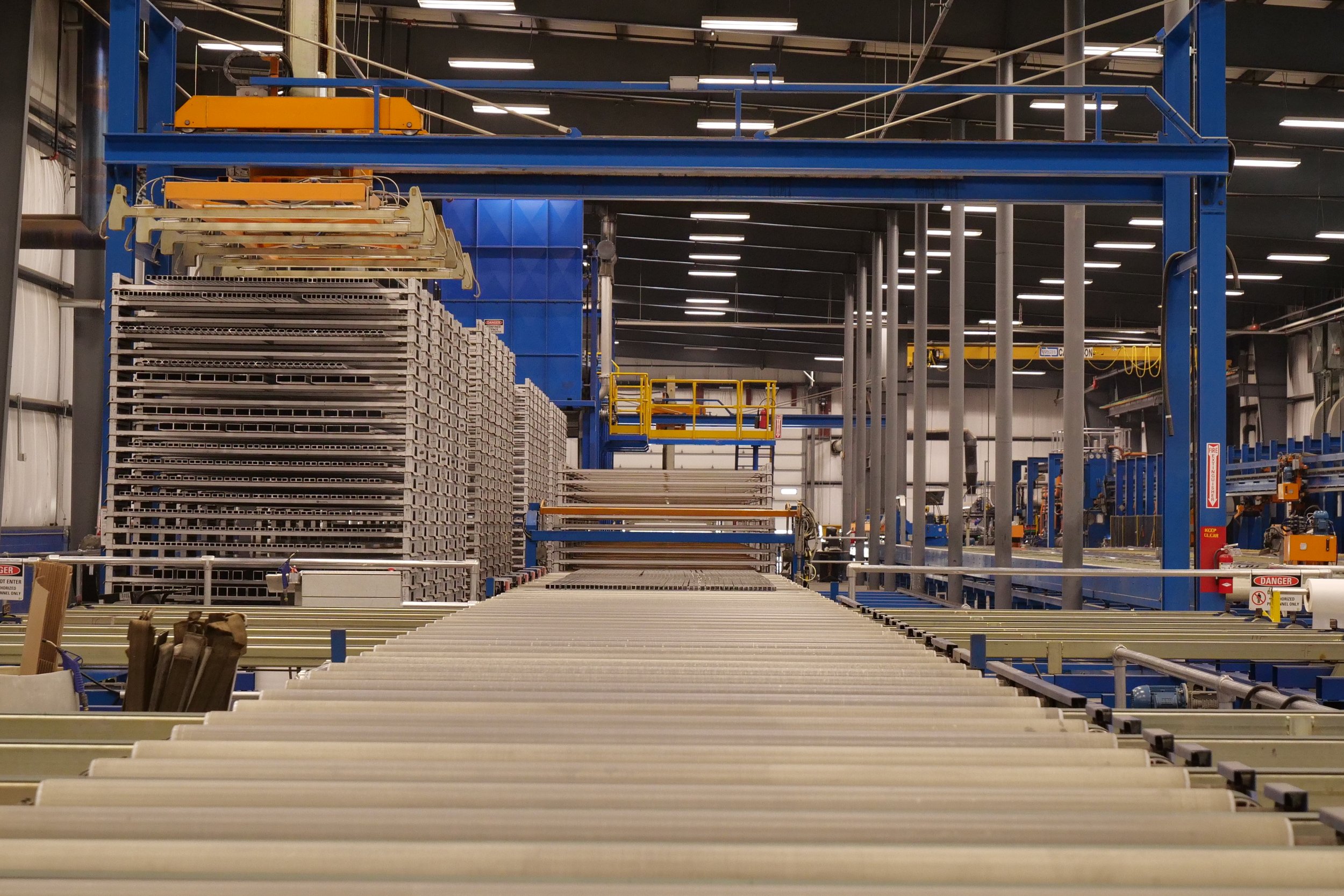 Industrial warehouse with metal racks and conveyor system, stacks of metal frames, and yellow safety equipment.