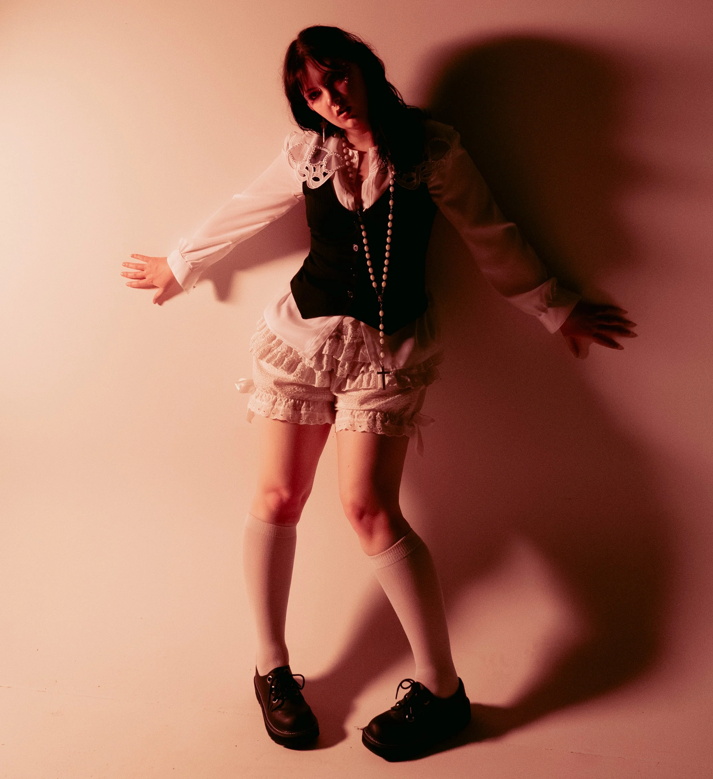 A woman wearing black combat boots, pink knee-high socks, a black vest, a white shirt with embroidered shoulders, a layered ruffled skirt, and a long bead necklace with a cross pendant, posing against a plain wall with shadows.