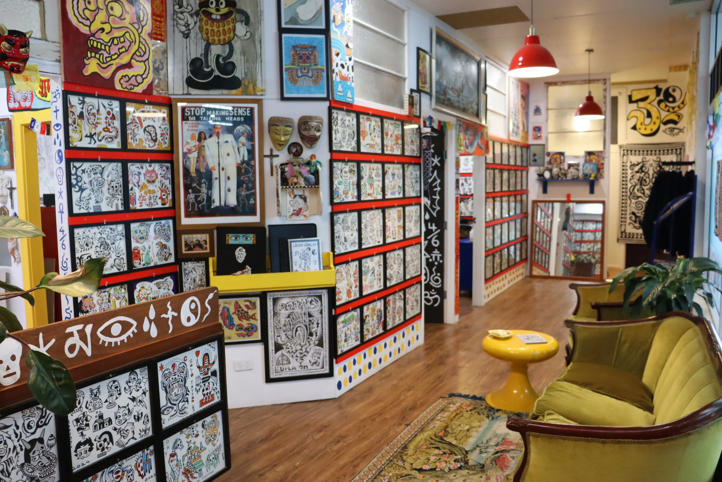 An art gallery with colorful artwork, masks, and framed pictures displayed on walls. There are yellow and green vintage armchairs, a small yellow table, and a patterned rug on wooden floors. Red pendant lights hang from the ceiling, and potted plants are present. The space has a cozy, eclectic vibe with a variety of art styles.