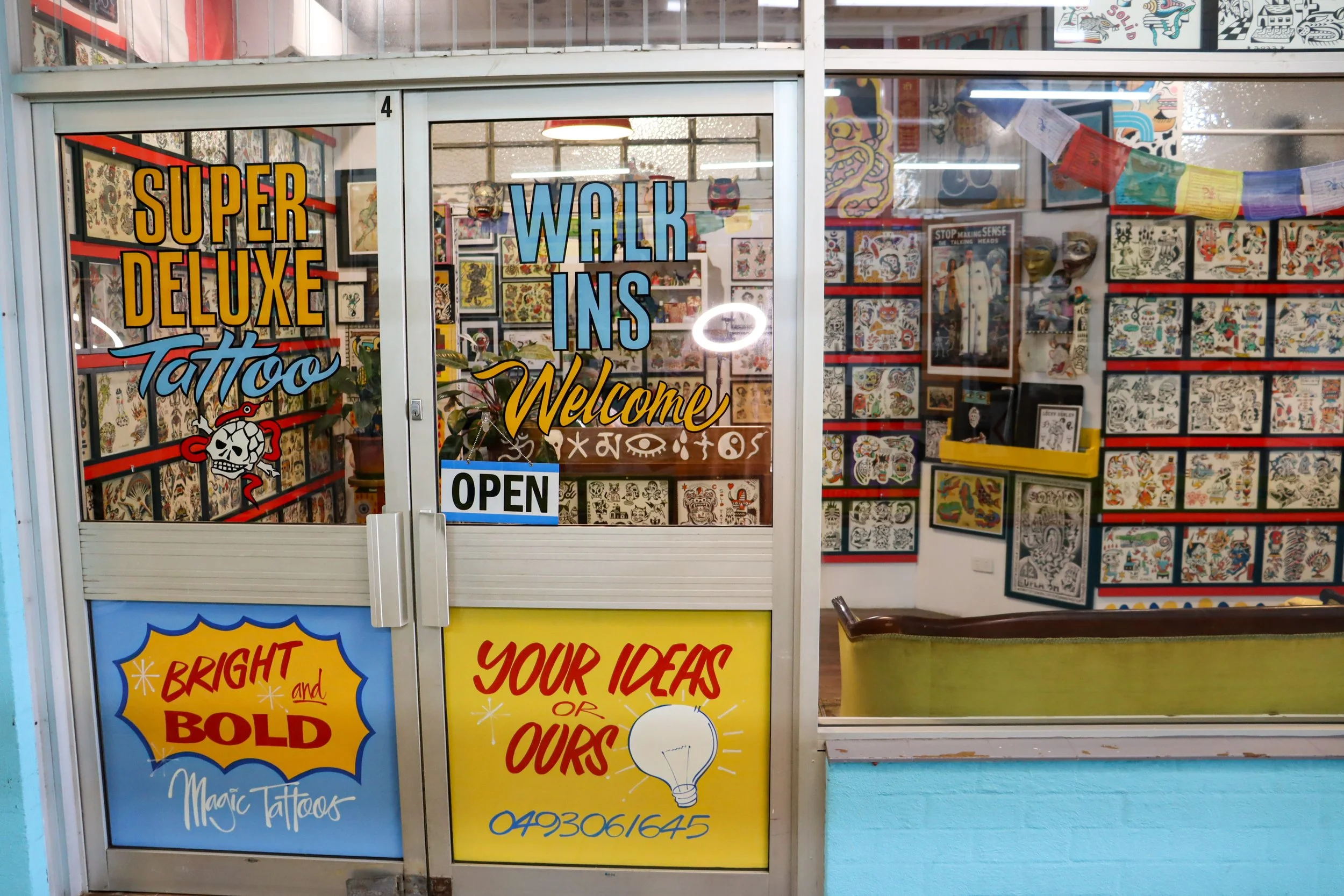 Tattoo shop front with colorful signs and framed tattoo artwork on the walls inside. The shop offers super deluxe tattoos, welcomes walk-ins, and is currently open. The door features bright posters promoting bold, creative tattoo ideas with a contact number.