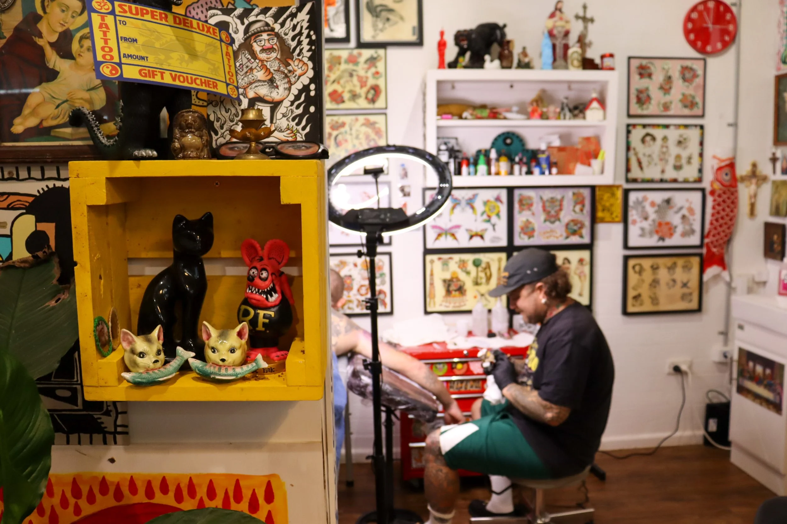An art studio with various tattoo designs on wall, a person getting a tattoo, and tattoo supplies. Tattoos include flowers and animals, and artwork adorns the walls.