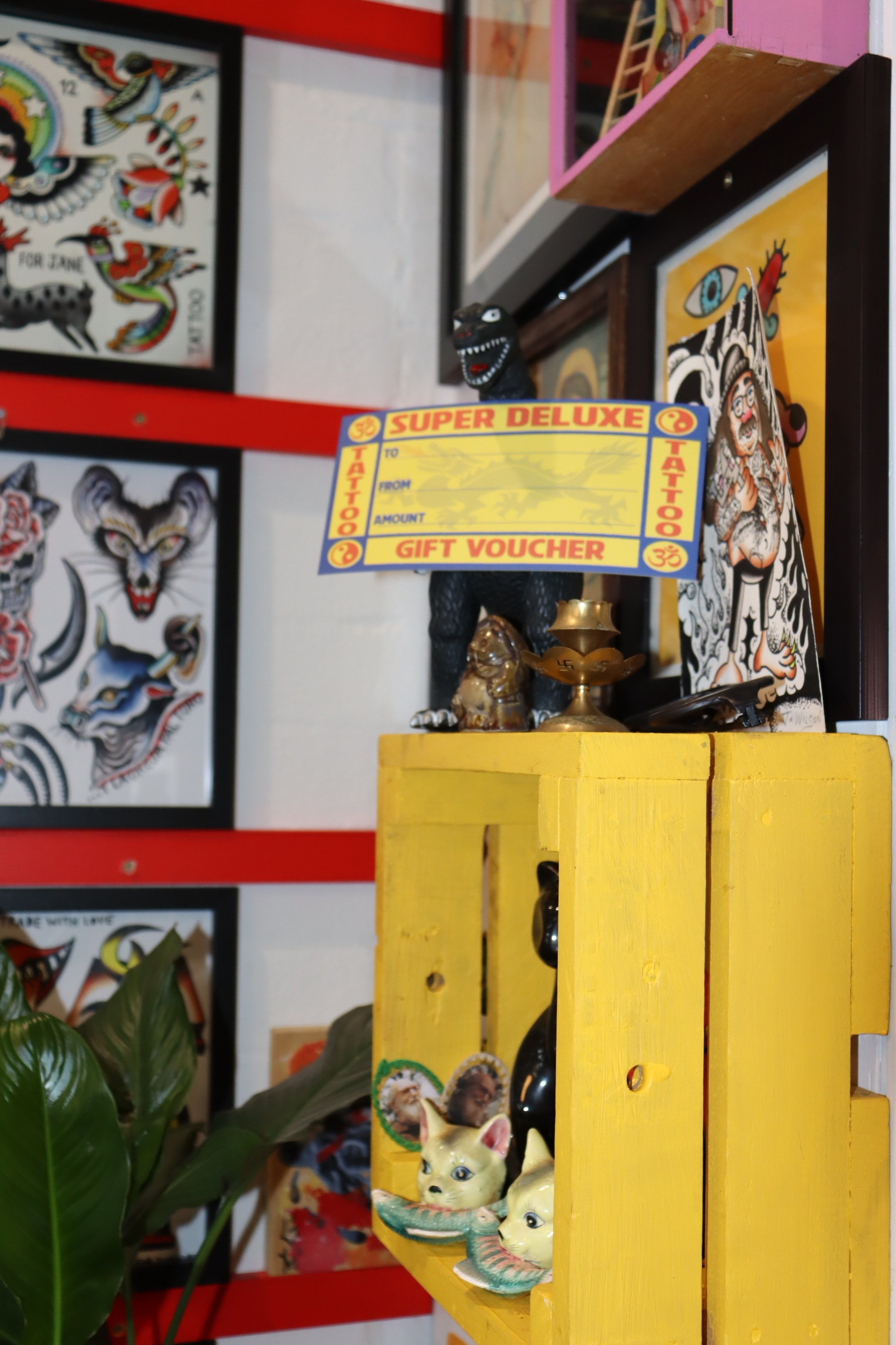 Decorative tattoo art and Asian-inspired figurines displayed on a yellow wooden shelf, with tattoo designs framed on the wall in the background.