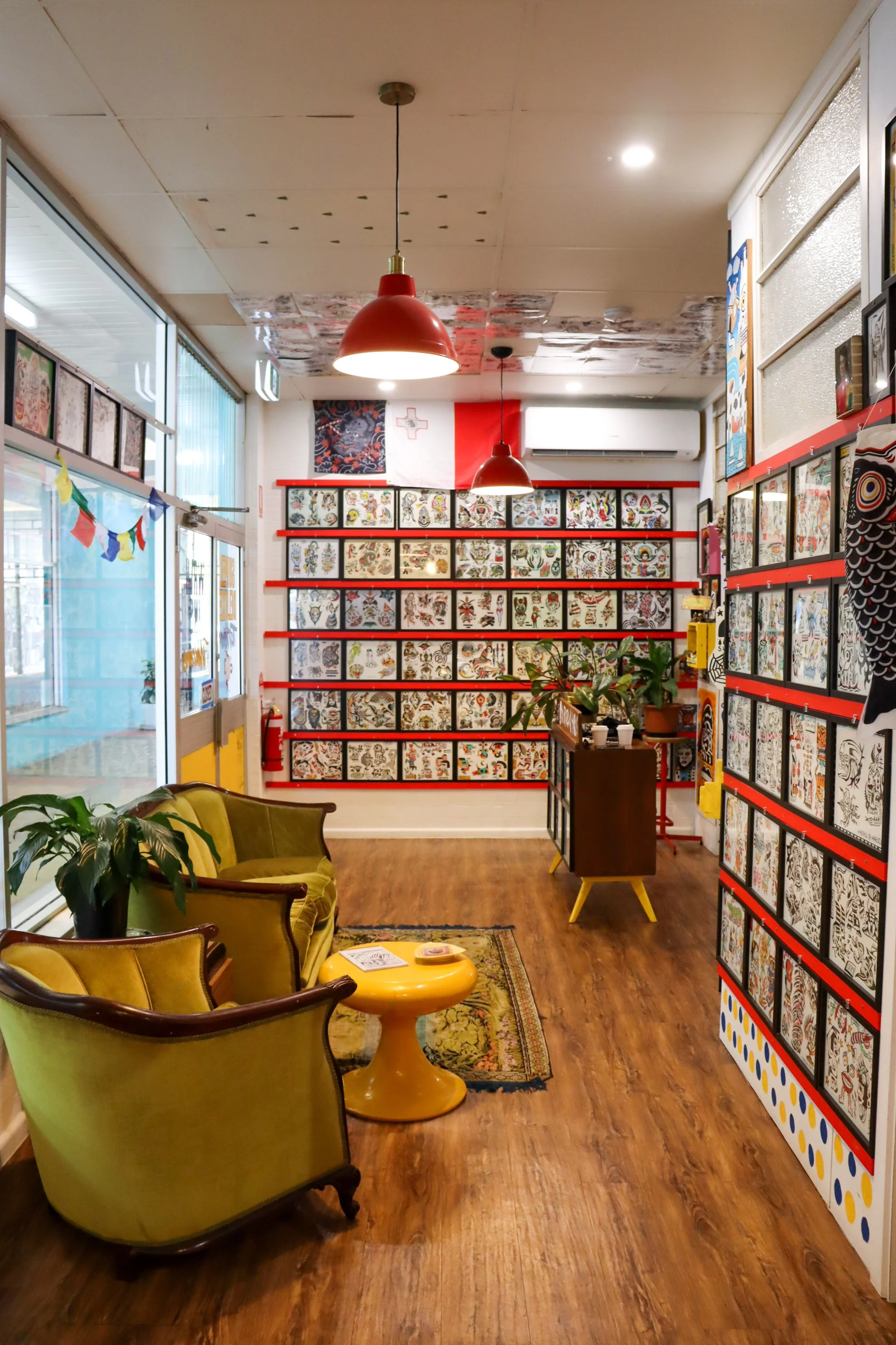 A cozy interior with wooden flooring, vintage green armchairs, yellow coffee table, indoor plants, red pendant lights, and walls decorated with framed tattoo-style artwork; a window on the left side and flags hanging near the door.