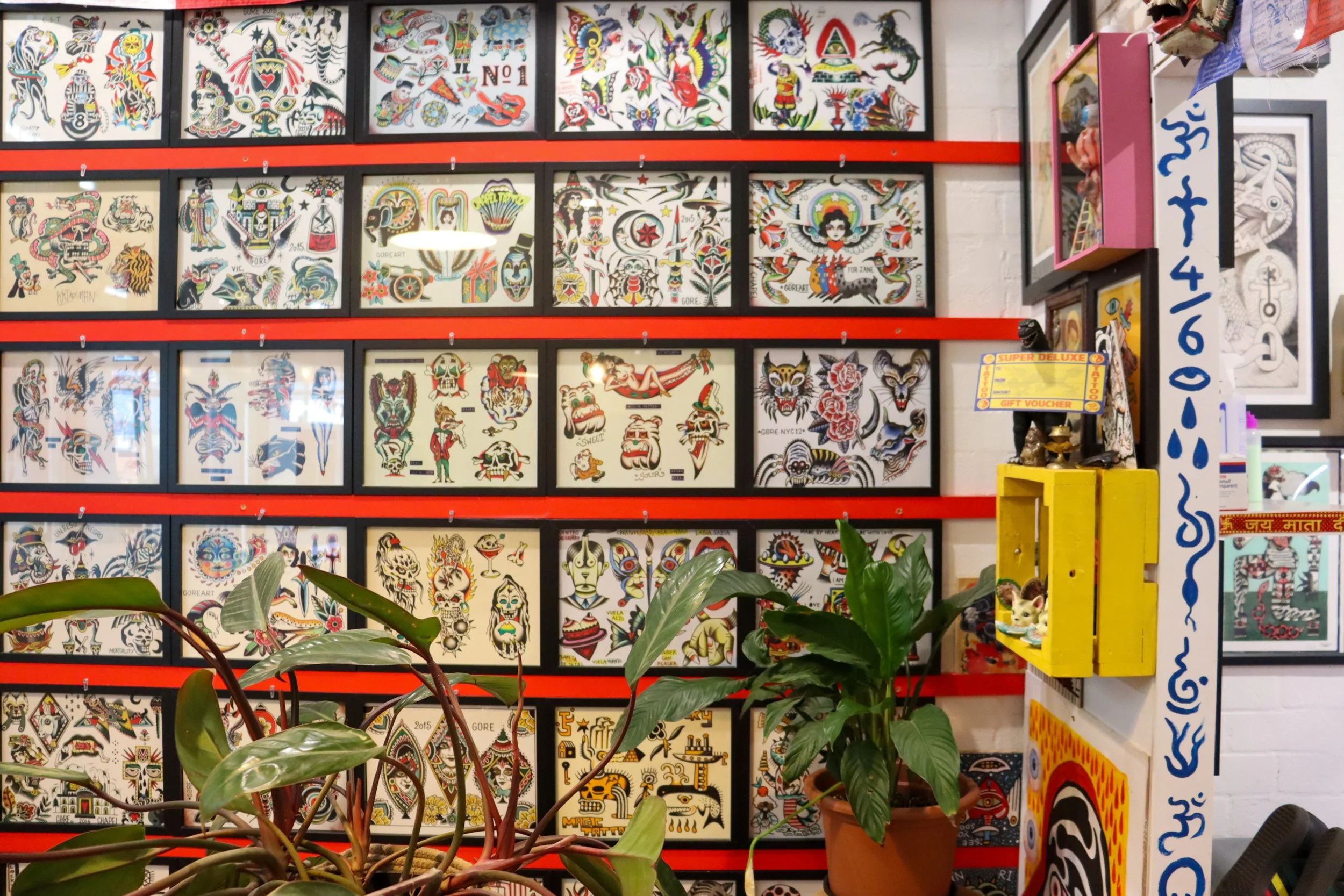 Wall decorated with framed traditional and colorful tattoo-style art, potted plants in front, and small shelves with decorative items on the right.