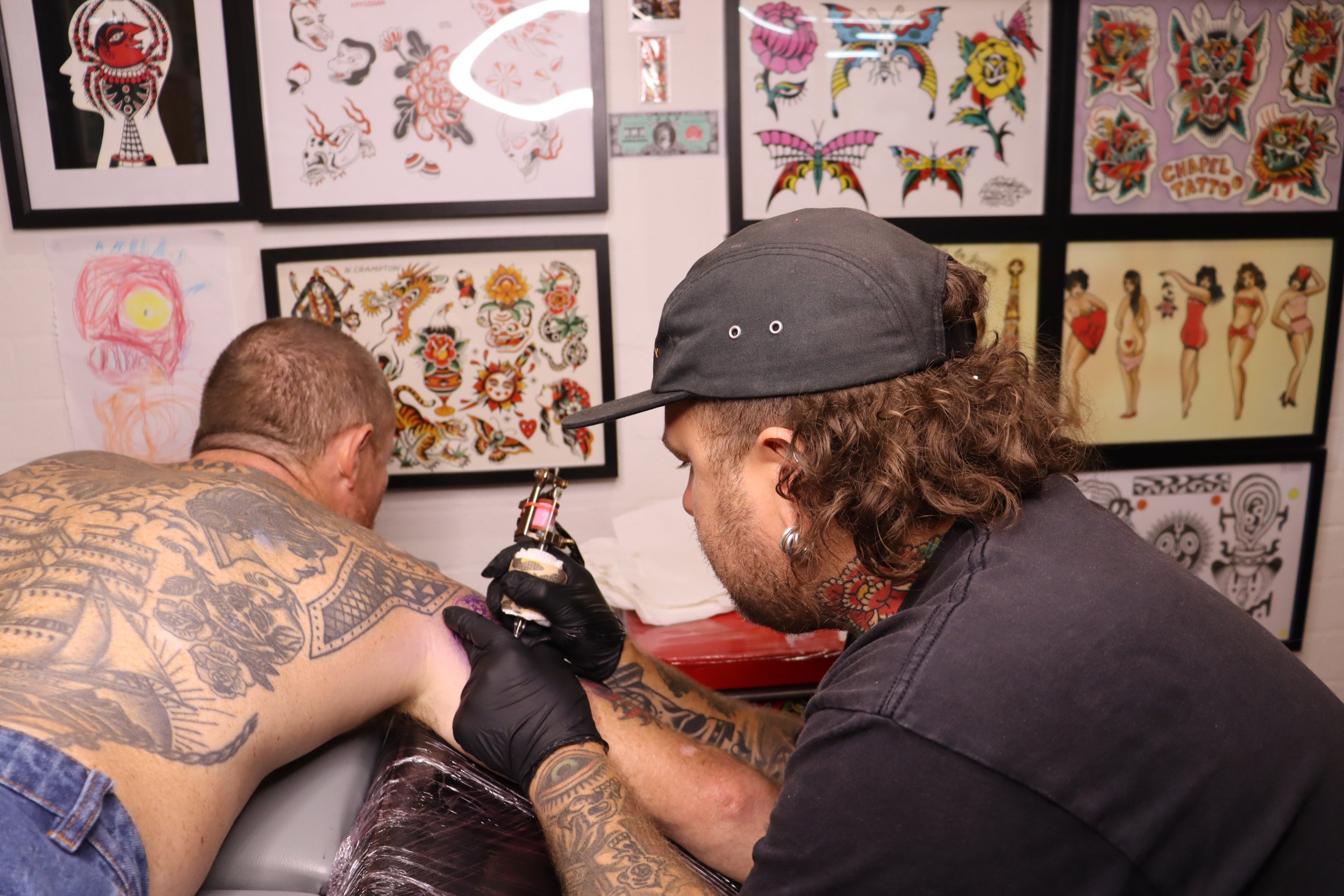 A man with tattoos receiving a tattoo on his shoulder from an artist in a tattoo studio decorated with framed tattoo designs and artwork.