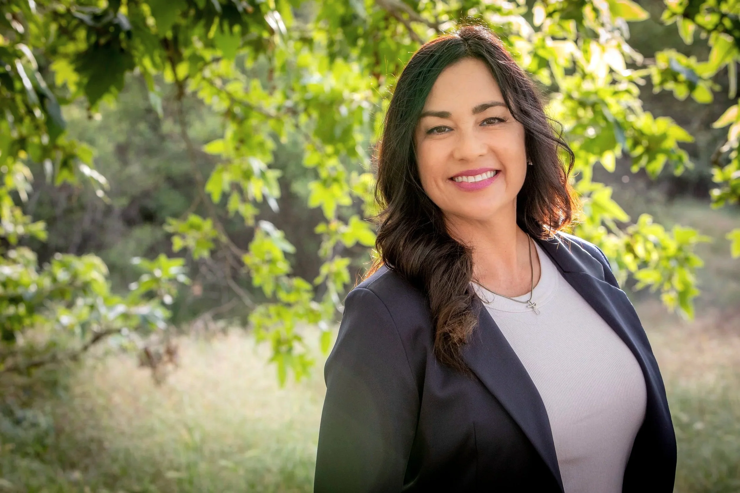 A smiling woman with long dark hair, wearing a red blouse and a dark blazer, standing outdoors in front of a building.