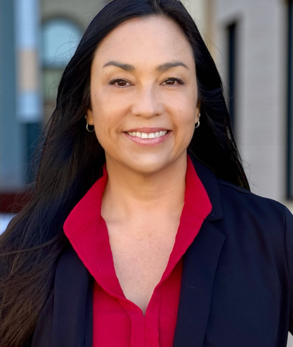 A smiling woman with long dark hair, wearing a red blouse and a dark blazer, standing outdoors in front of a building.