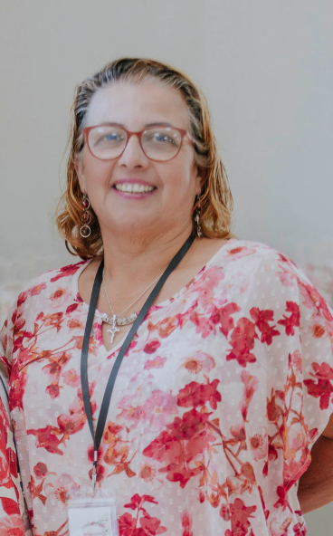 A woman with glasses and shoulder-length hair smiling, wearing a pink floral blouse and a lanyard.