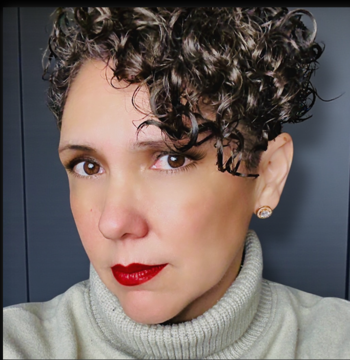 Close-up of a woman with short, curly hair, red lipstick, wearing a beige turtleneck sweater and diamond earrings.