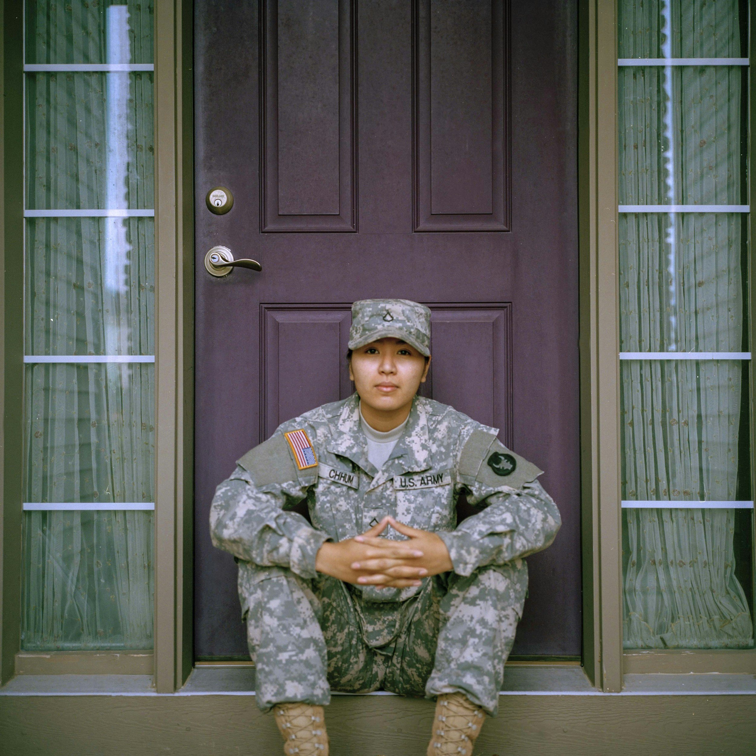Young woman in military uniform sitting with legs crossed in front of a purple door, with her hands clasped together, wearing a camouflage hat and boots.