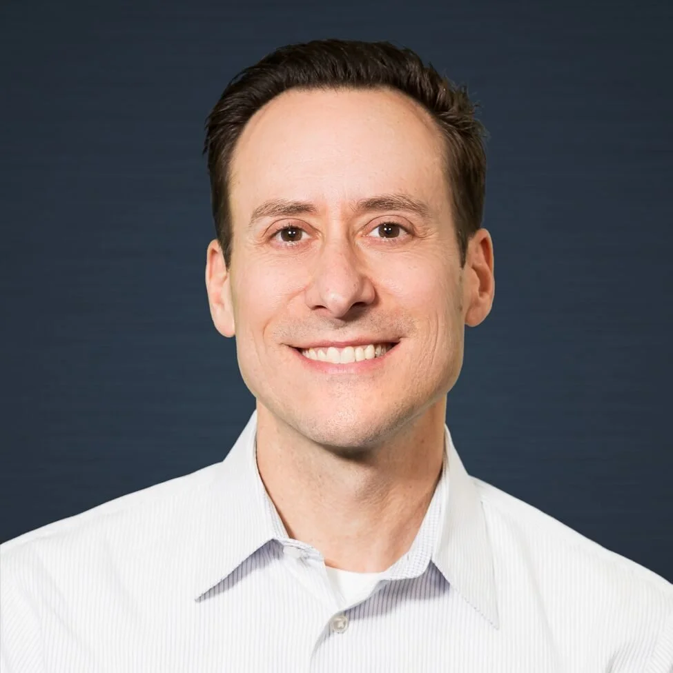 Headshot of a man with short dark hair, smiling, in a light-colored collared shirt against a dark gray background.
