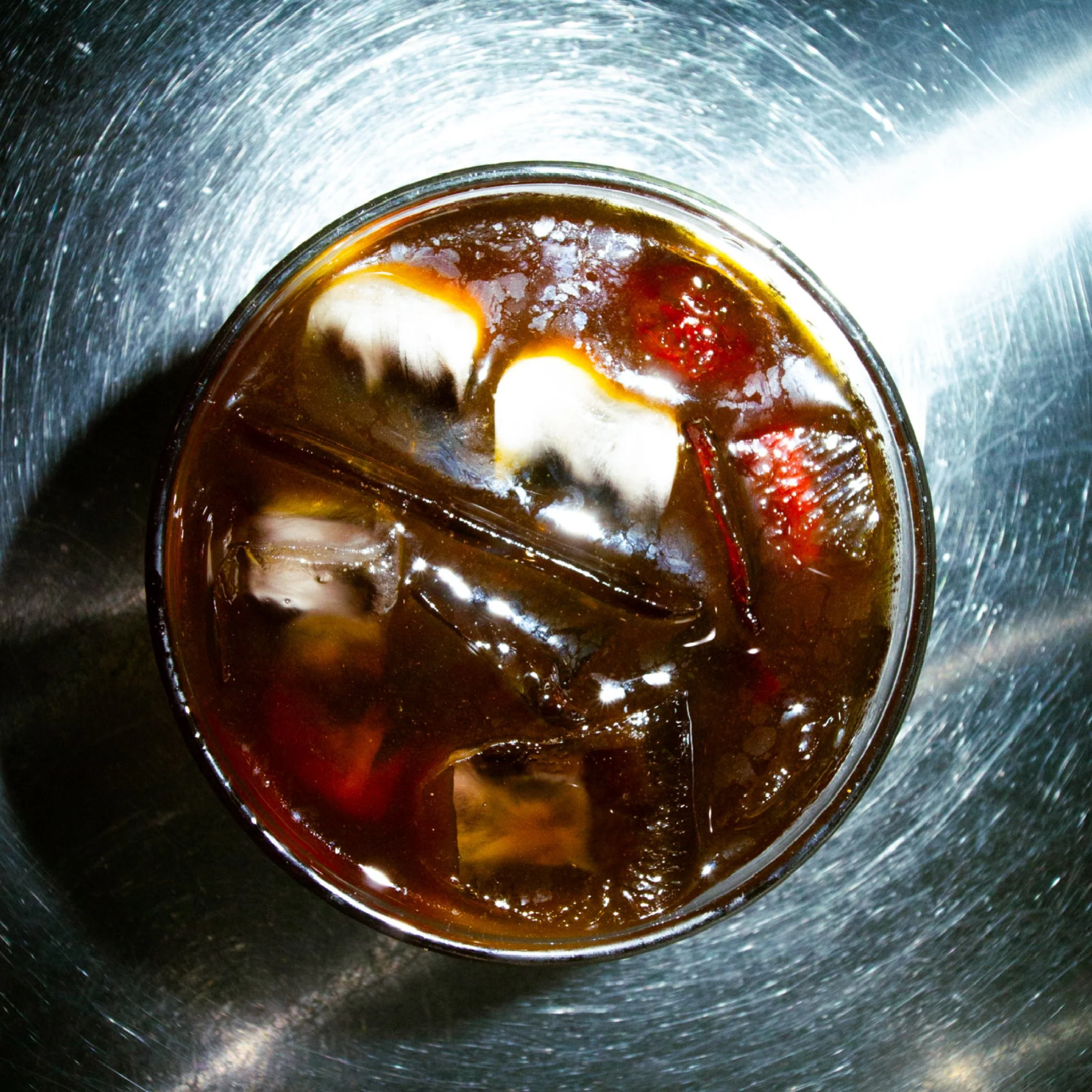 A top-down view of a glass filled with a dark soda and ice cubes on a shiny metal surface.