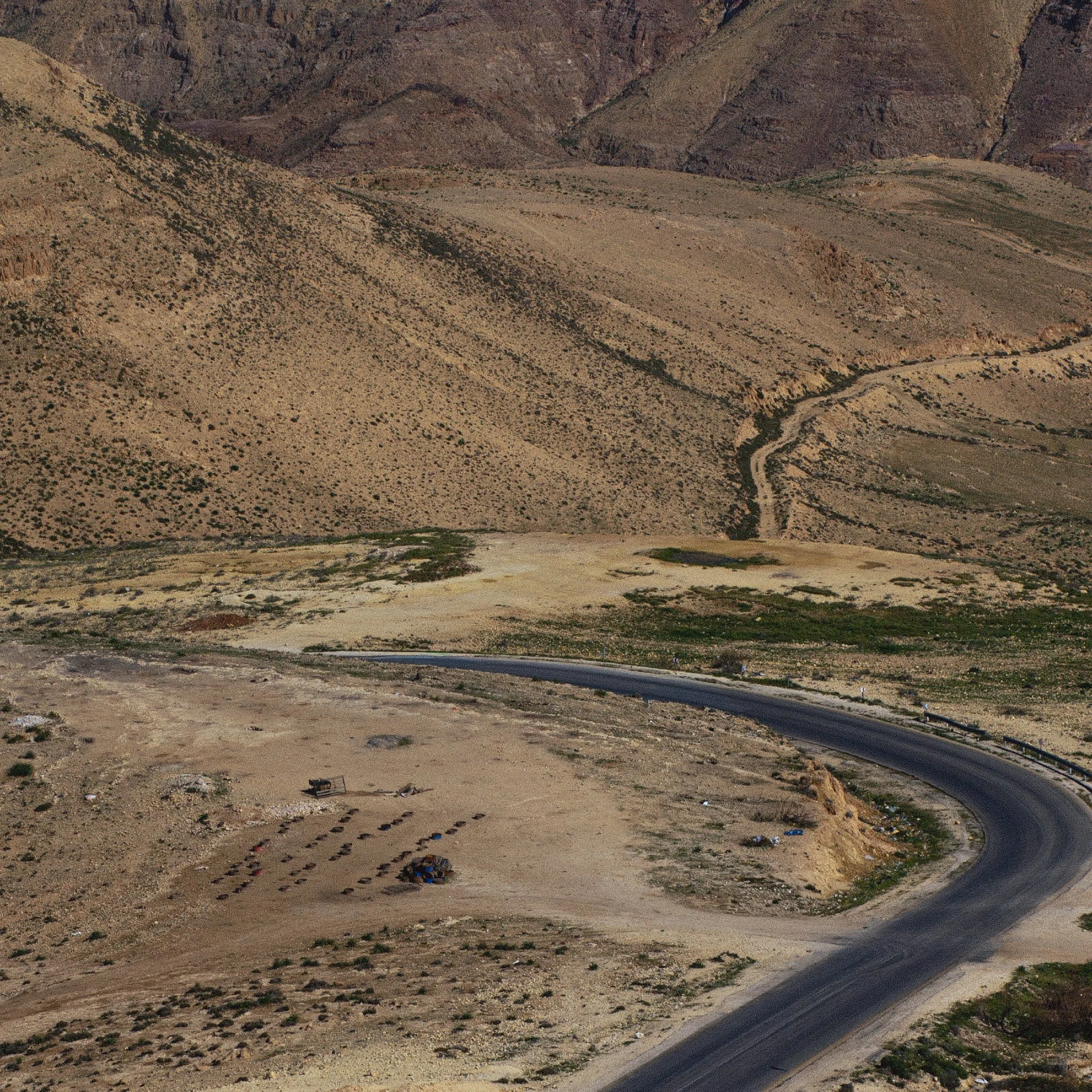 A winding mountain road through a dry, rugged landscape with sparse vegetation and distant mountain peaks.