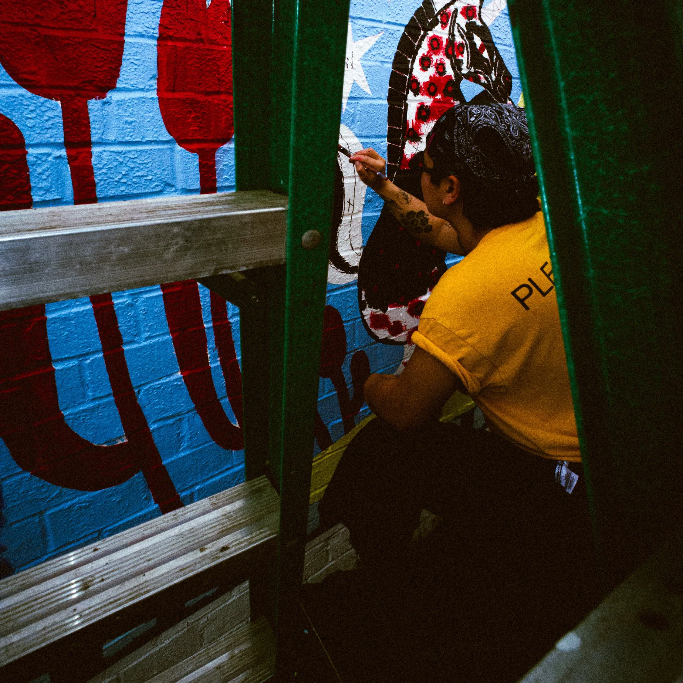 A person with a bandana and glasses, wearing a yellow shirt, is painting a mural on a brick wall that features a face with long hair and glasses, in front of a red and blue background.