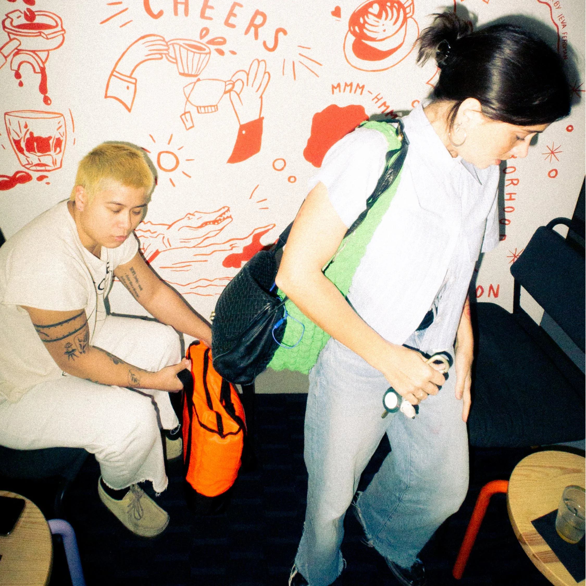 A person with dark hair and glasses is standing with their hands on their hips, while another person with bleached blonde hair and tattoos is sitting behind them and holding an orange bag. The background features red illustrations and text about chee