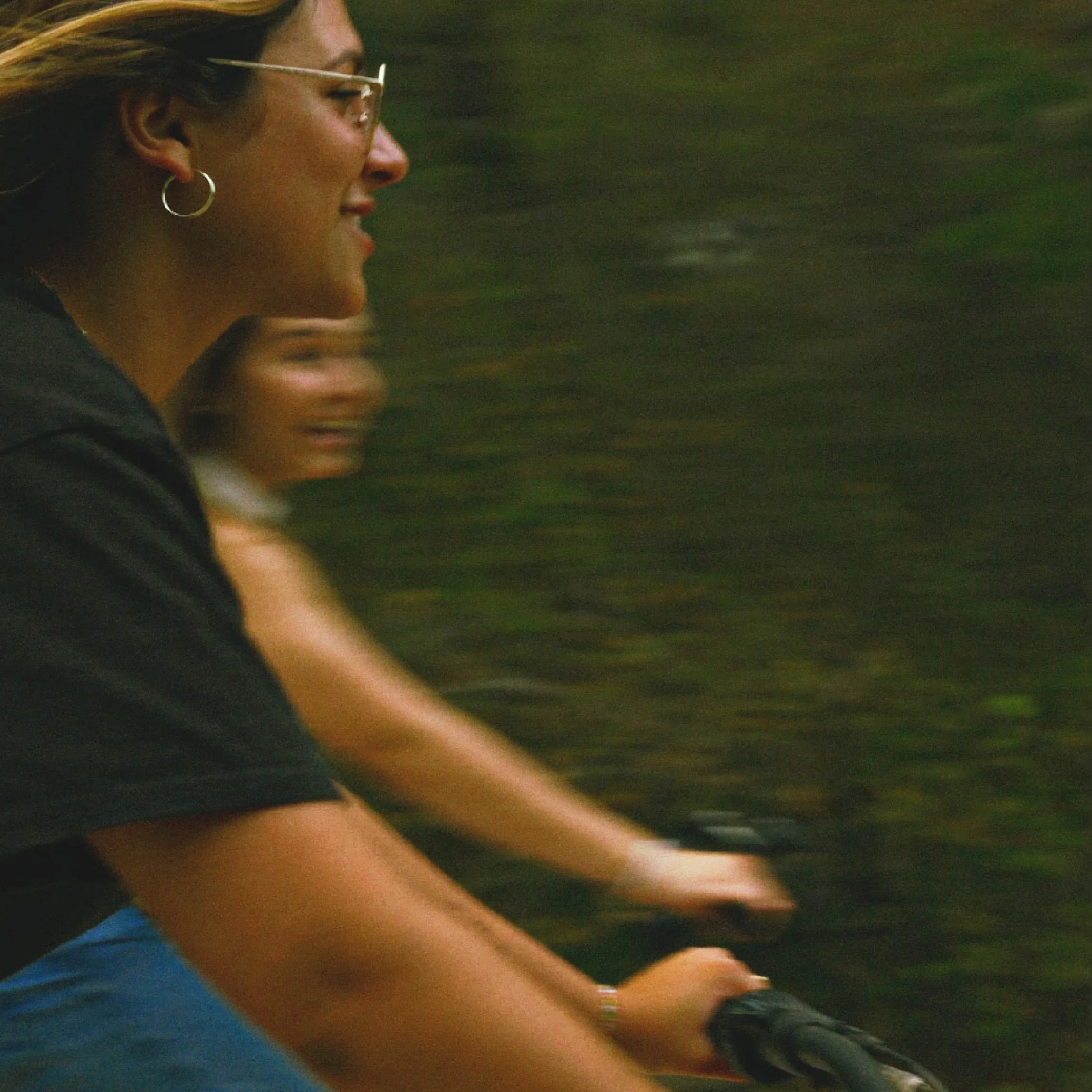 A woman with sunglasses and hoop earrings is riding a bicycle through a green outdoor area.