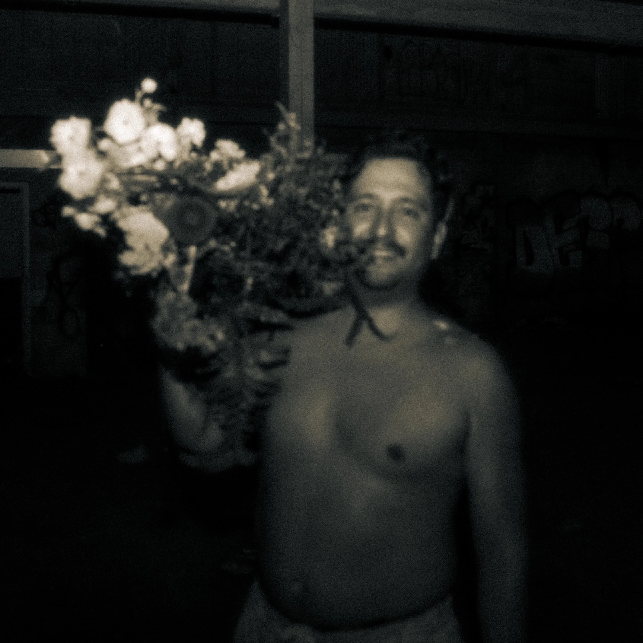 A shirtless man with a beard and mustache holding a large bouquet of flowers, smiling, with a graffiti-covered wall in the background.
