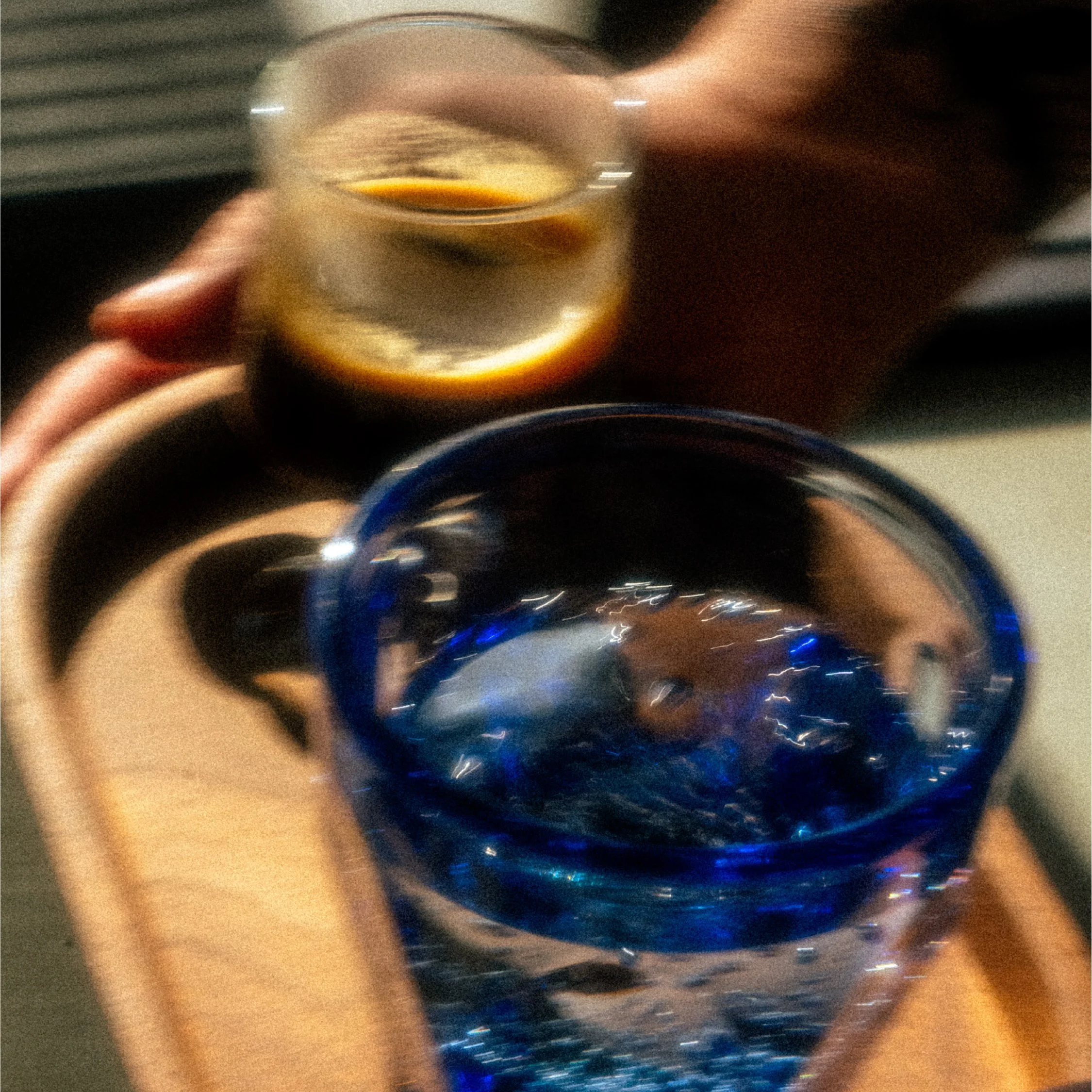 A hand holding a glass of dark beverage with ice, and another glass with a dark drink, both on a wooden tray.