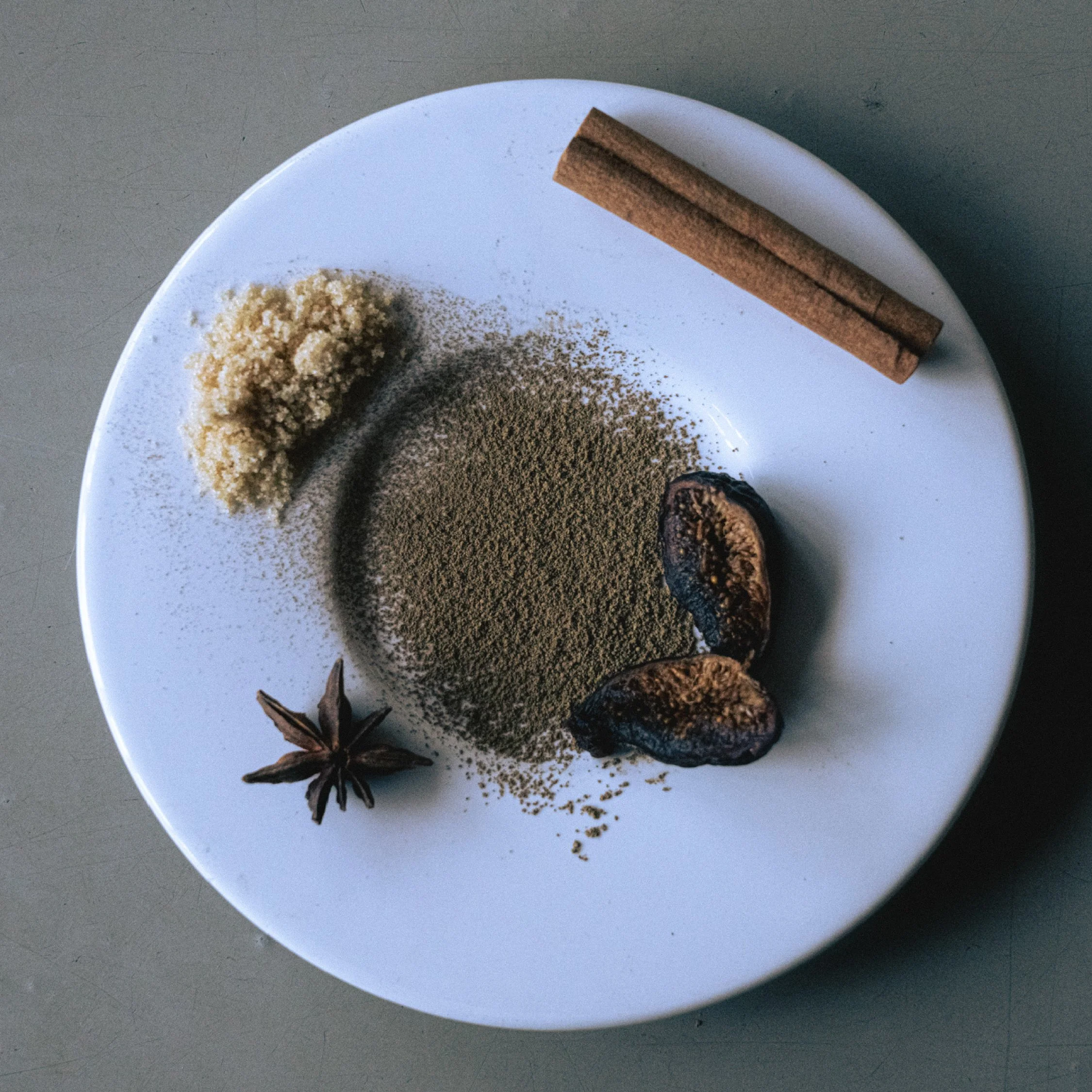 White plate with spices, including cinnamon sticks, star anise, dried ginger, ground pepper, and dried cardamom pods.