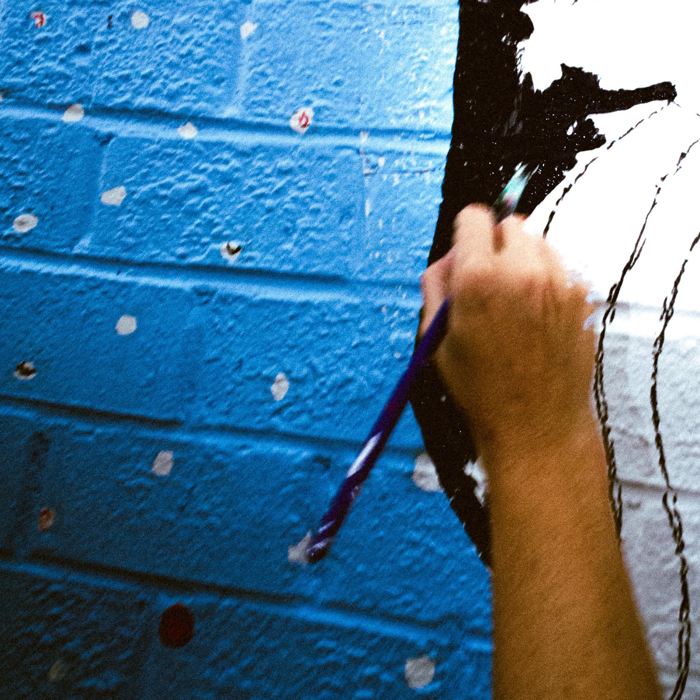 A person painting a mural on a blue brick wall with white and black paint, using a paintbrush.