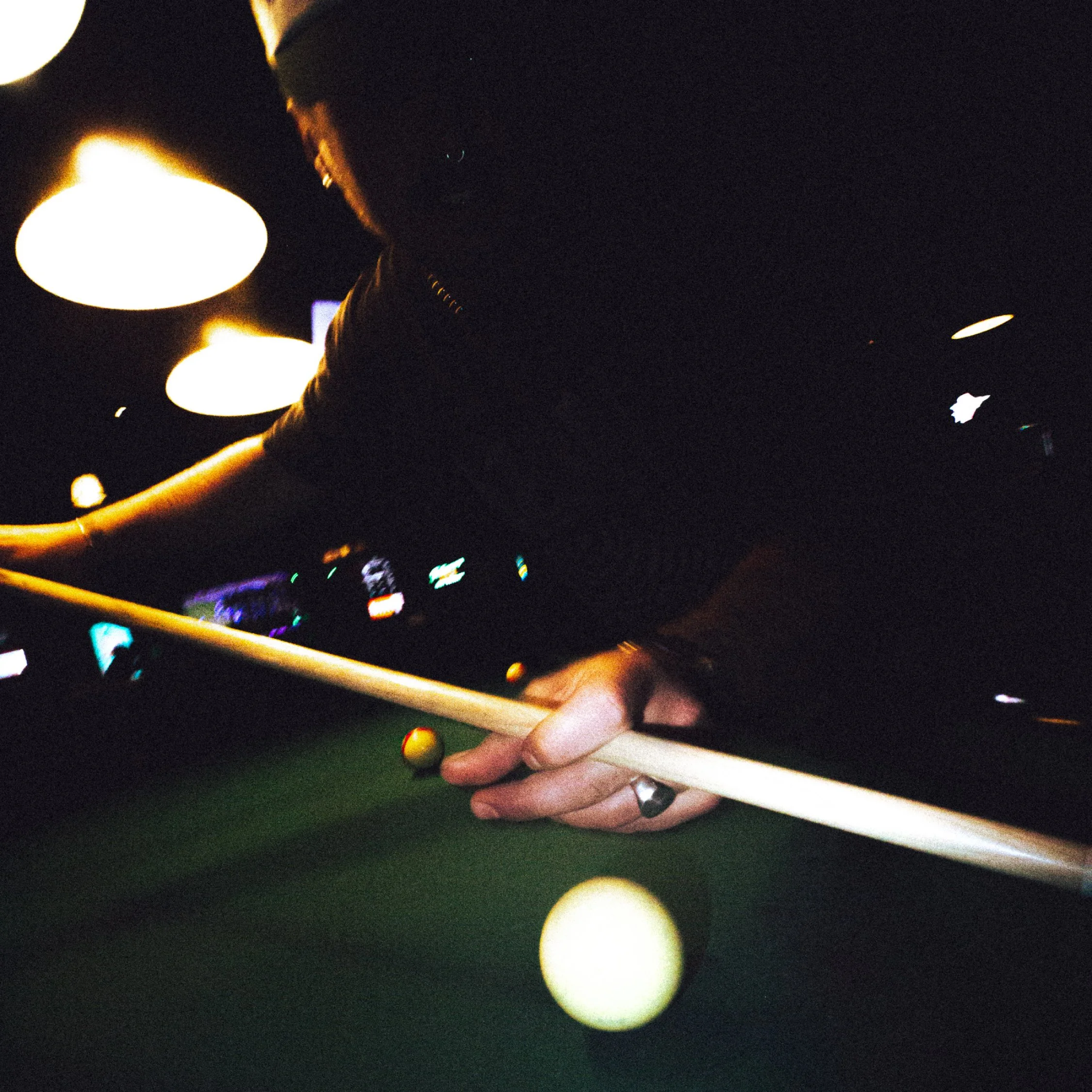 A person playing pool in a dimly lit room, focused on aiming the cue ball with a cue stick.