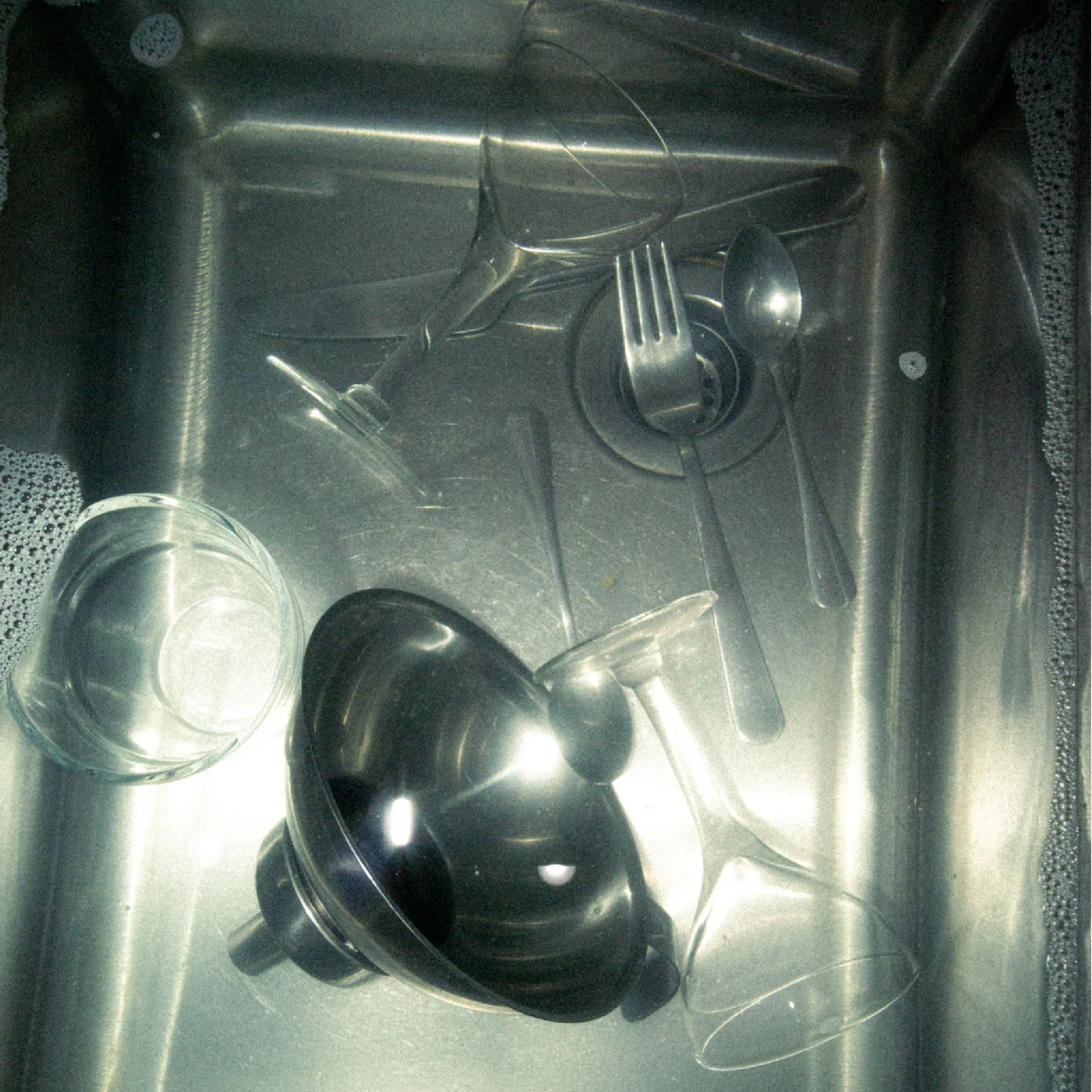 Dish drying area with various kitchenware including a glass, a metal strainer, a metal bowl, a spoon, a fork, and a metal cup, all placed on a stainless steel sink.