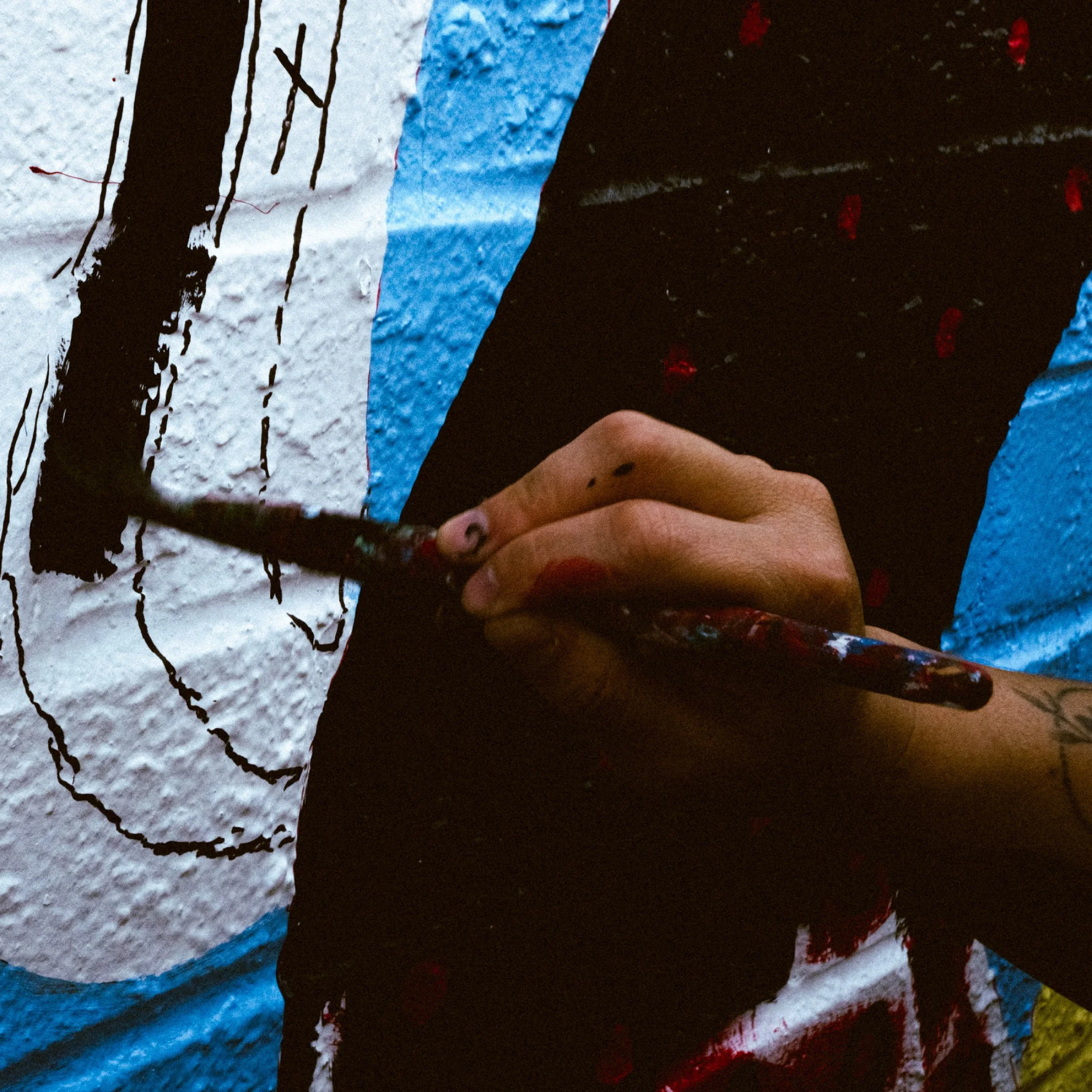 A person holding a paintbrush, painting black lines on a textured white wall with blue accents, part of a larger mural.