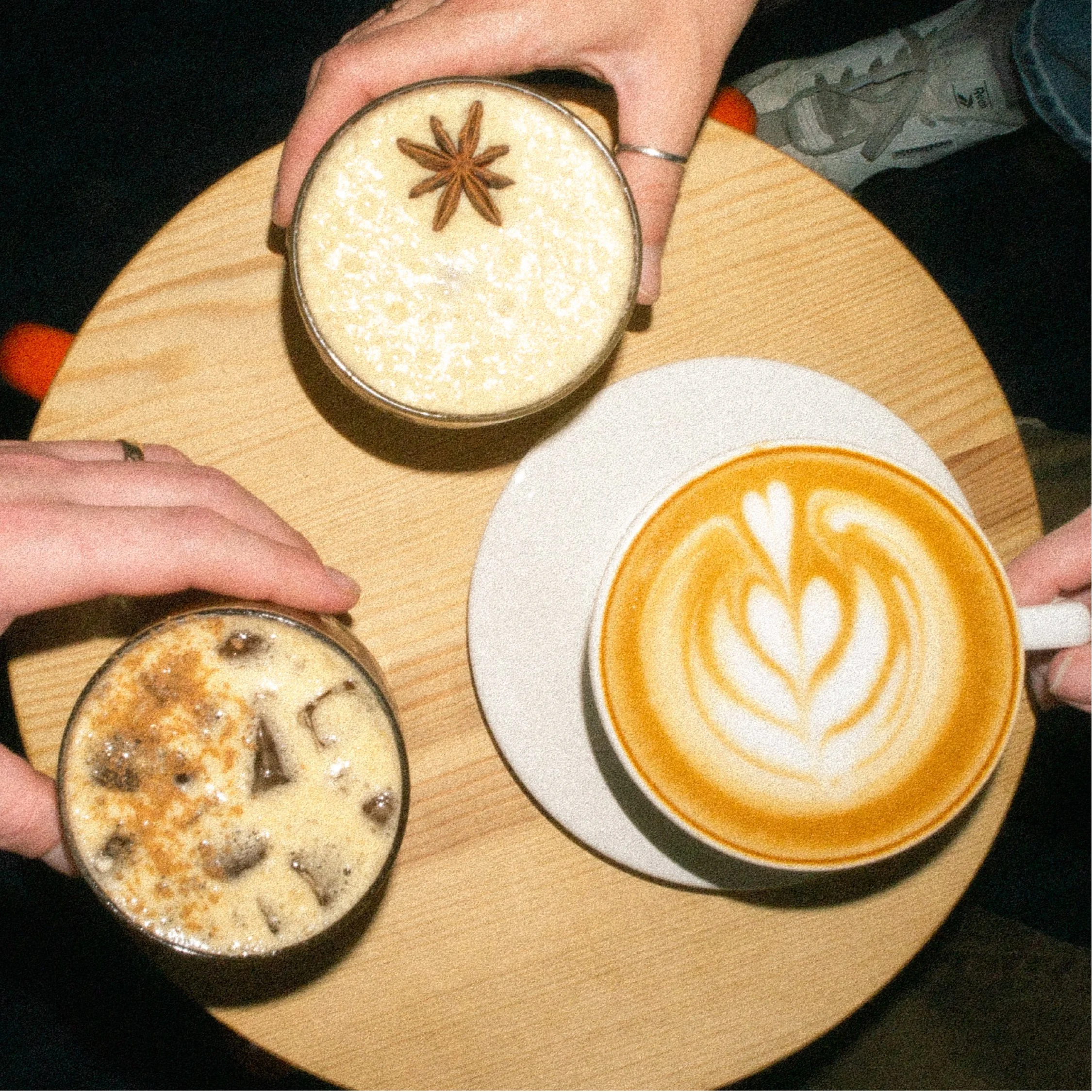 Two hands holding glasses of coffee or hot chocolate and a large cup of coffee with latte art on a round wooden table.