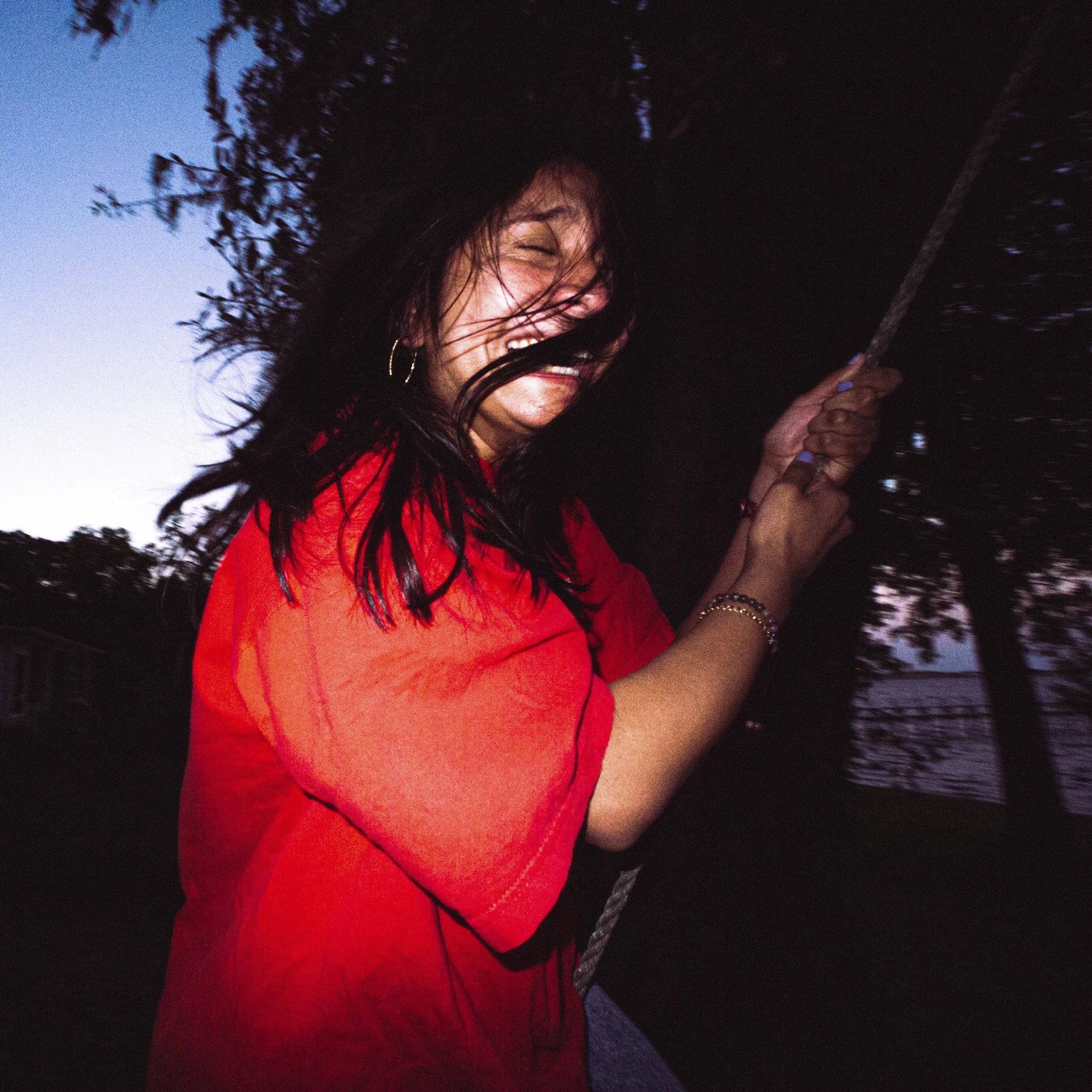 A woman with dark hair wearing a red shirt, smiling and holding onto a rope outdoors at dusk.