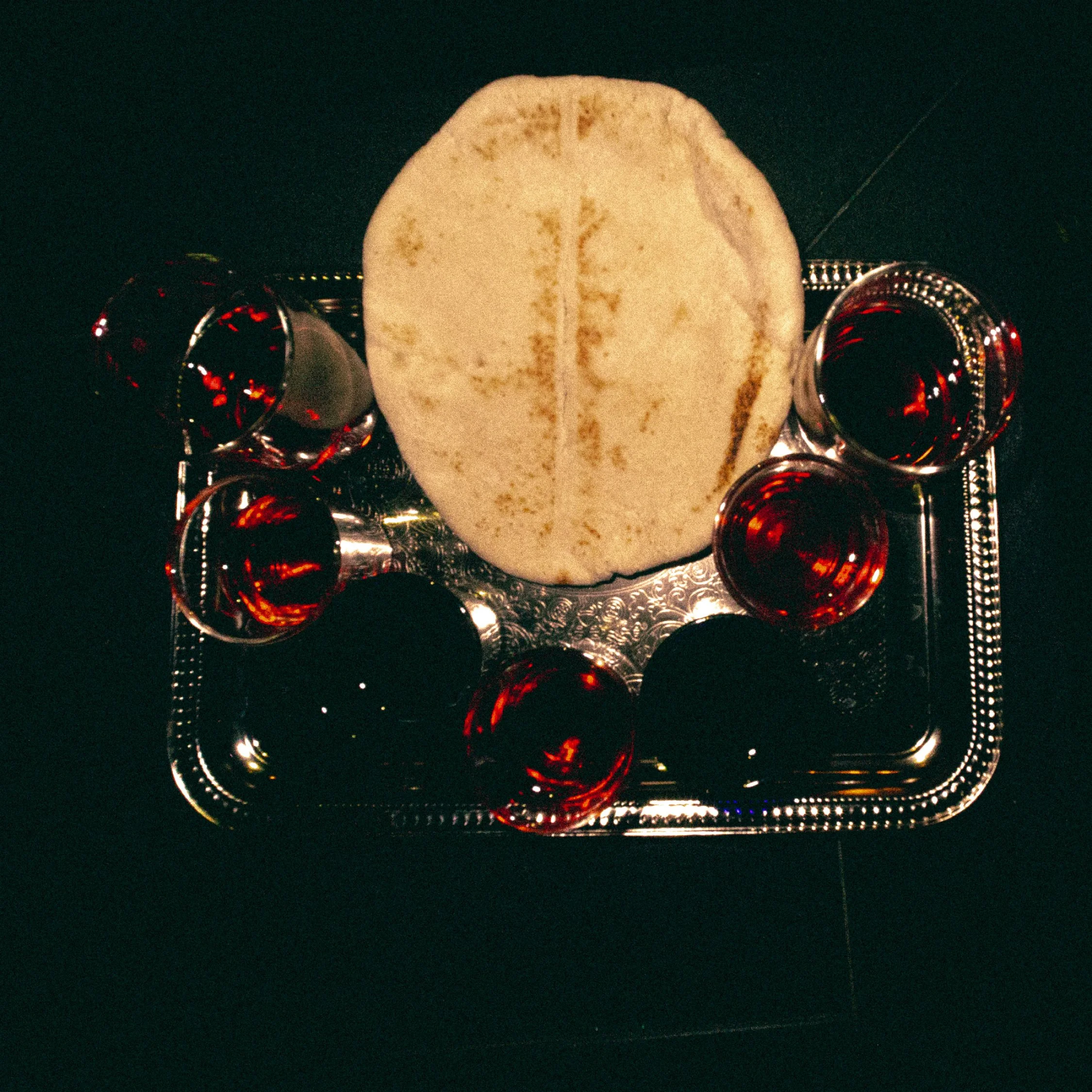 A silver tray with six glasses of red wine surrounding a piece of flatbread on a dark surface.