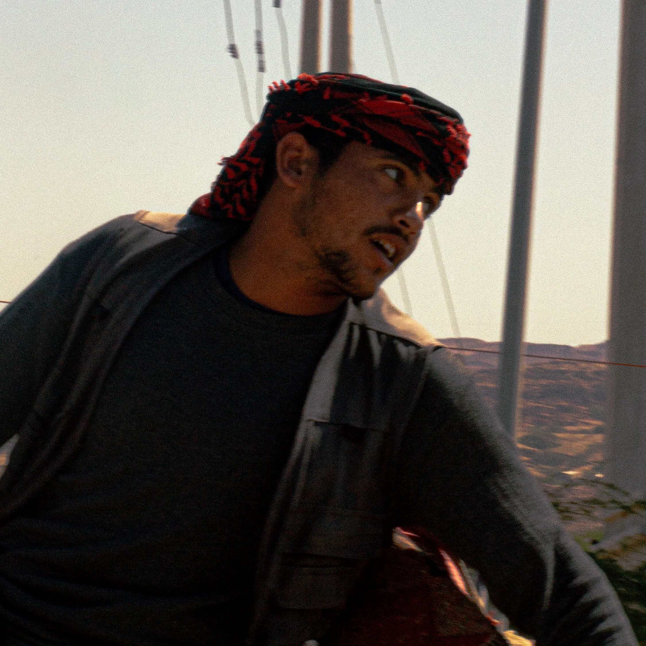 A man wearing a black shirt and a black leather jacket, with a red and black headscarf, leaning forward outdoors during sunset.