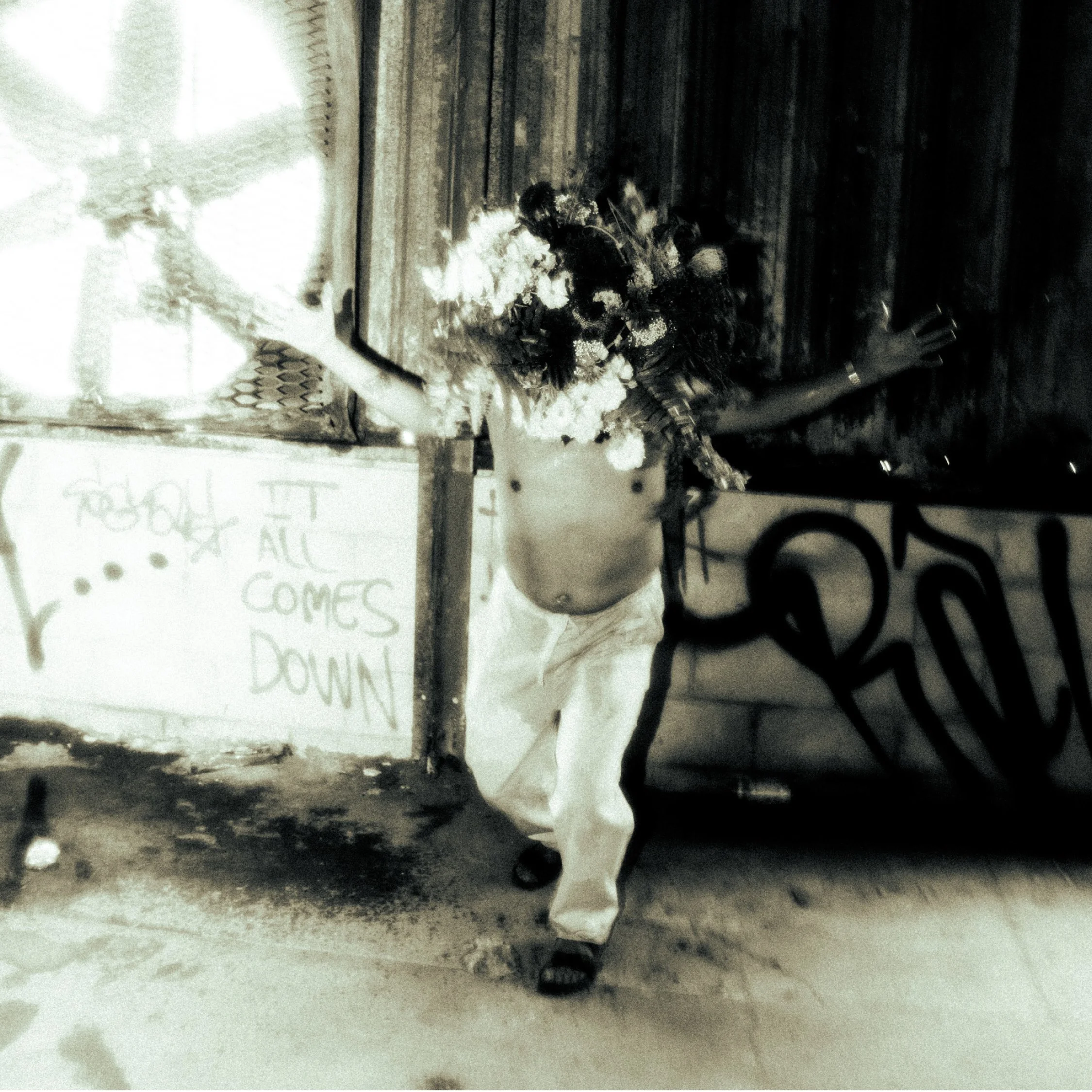 A man with a bare chest holding a large bouquet of flowers, standing in front of a wall with graffiti and a sign that reads "It all comes down." The image is in black and white.