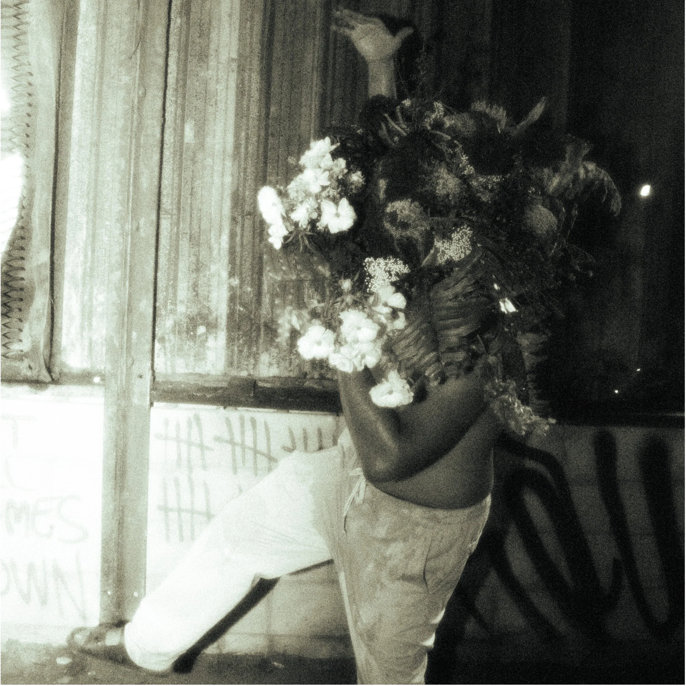 A shirtless man with a painted face and long hair, wearing light-colored pants, is bending down and holding a large bouquet of flowers.