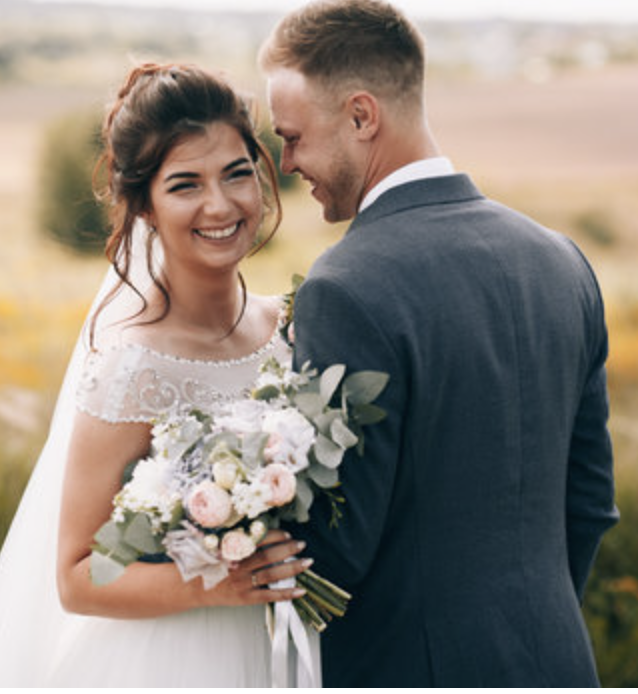 Smiling bride holding a bouquet of white and pink flowers, wearing a white wedding dress, standing close to groom in a blue suit outdoors in a field.