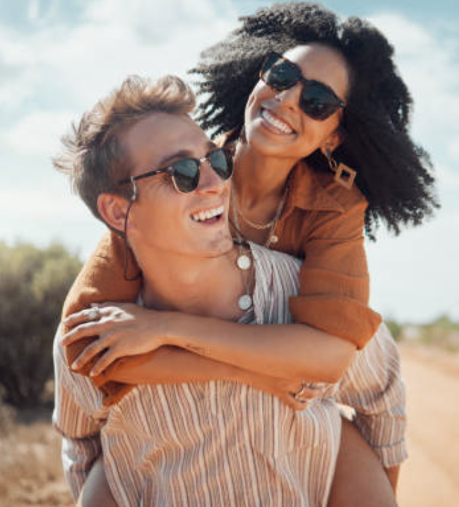 A smiling couple outdoors, the woman piggybacking on the man's back on a sunny day.