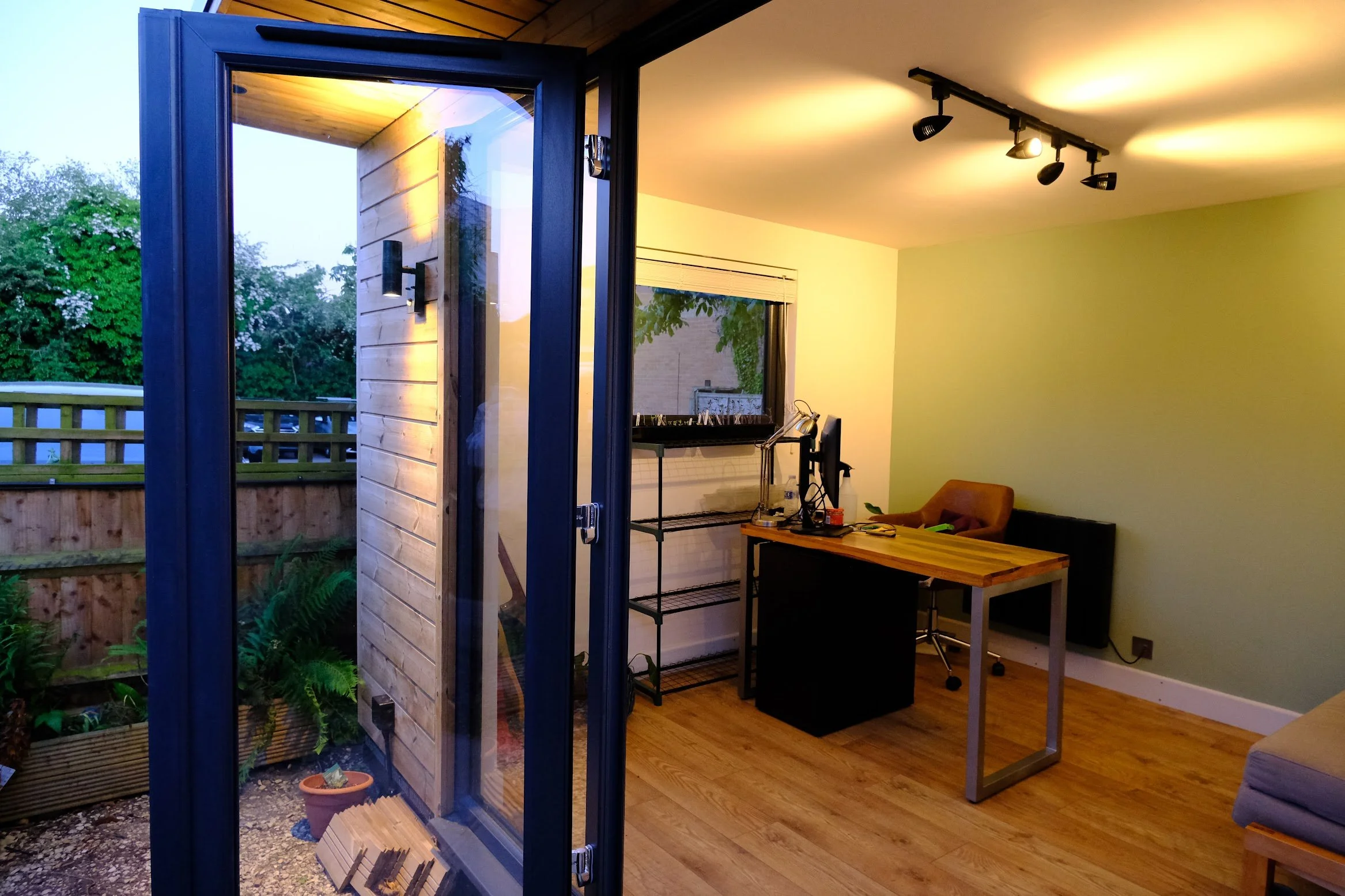 View of a home office from outside with an open glass door, showing a desk with a computer, a chair, a plant, and a window, illuminated by ceiling track lights, with greenery and a wooden fence visible outside.