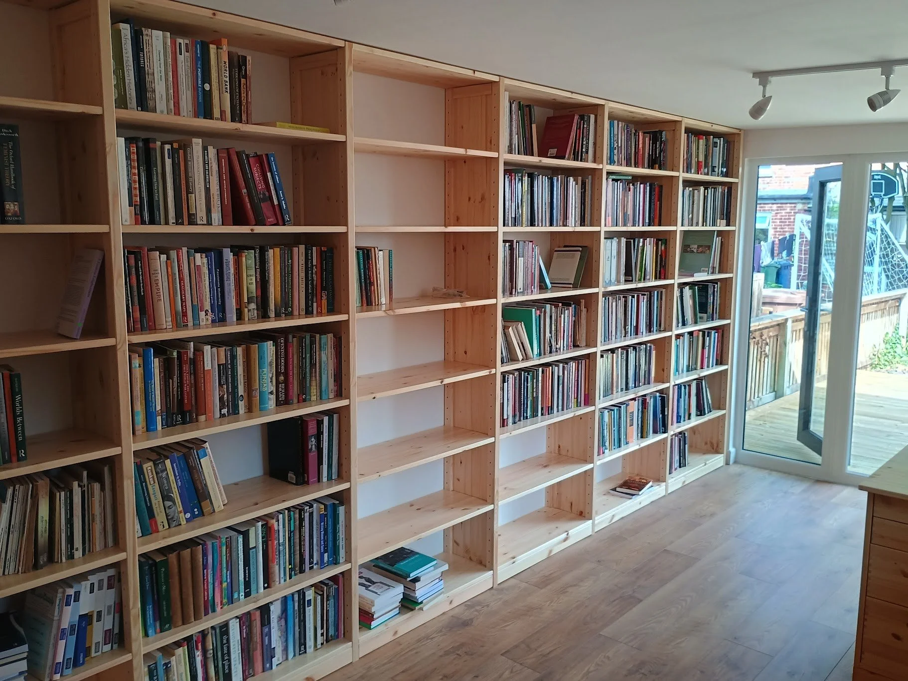 Empty wooden bookshelves filled with books in a room with hardwood flooring and sliding glass doors leading to an outdoor deck.